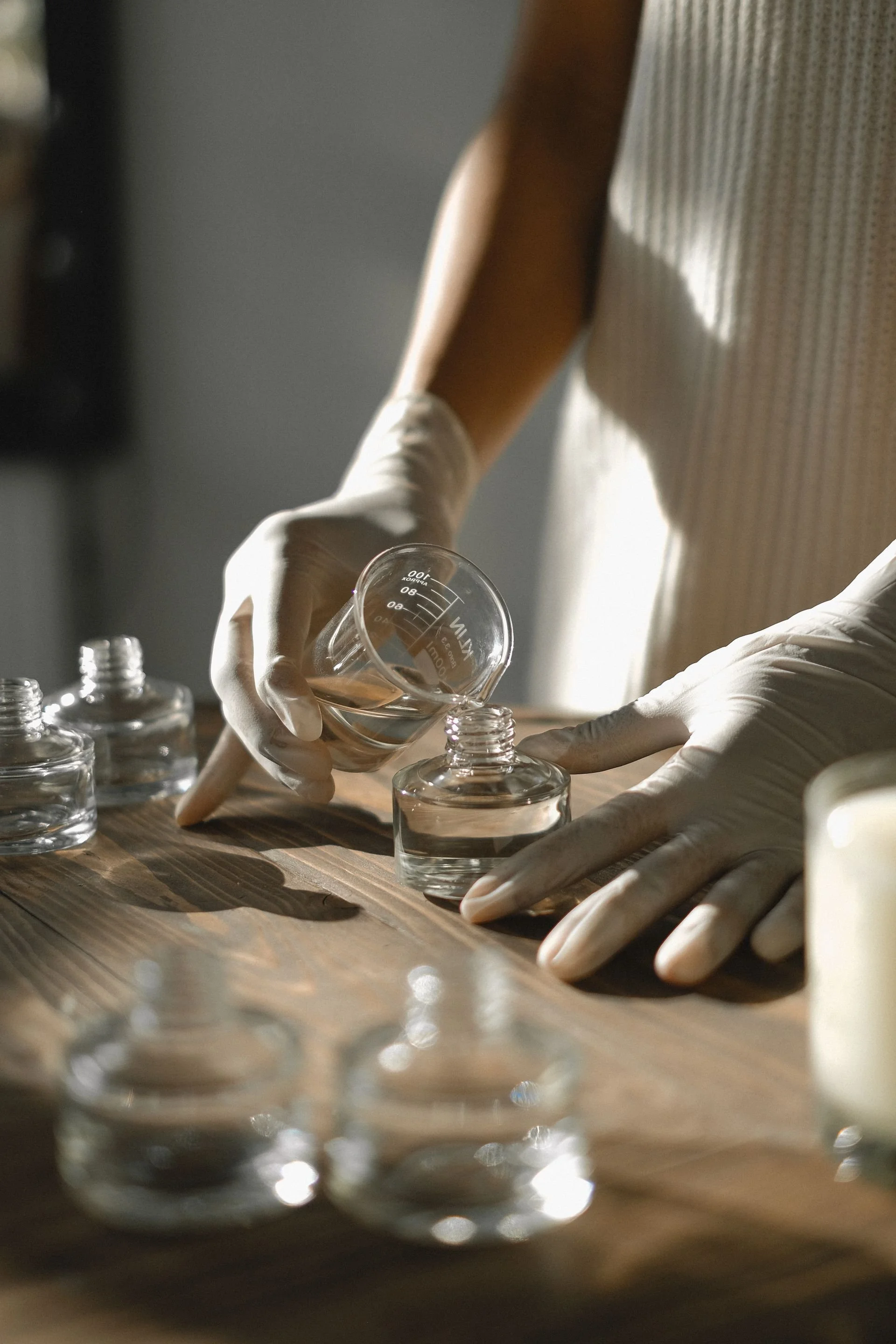 Gloved hands pouring a clear liquid from a glass beaker into small glass bottles on a wooden work surface.