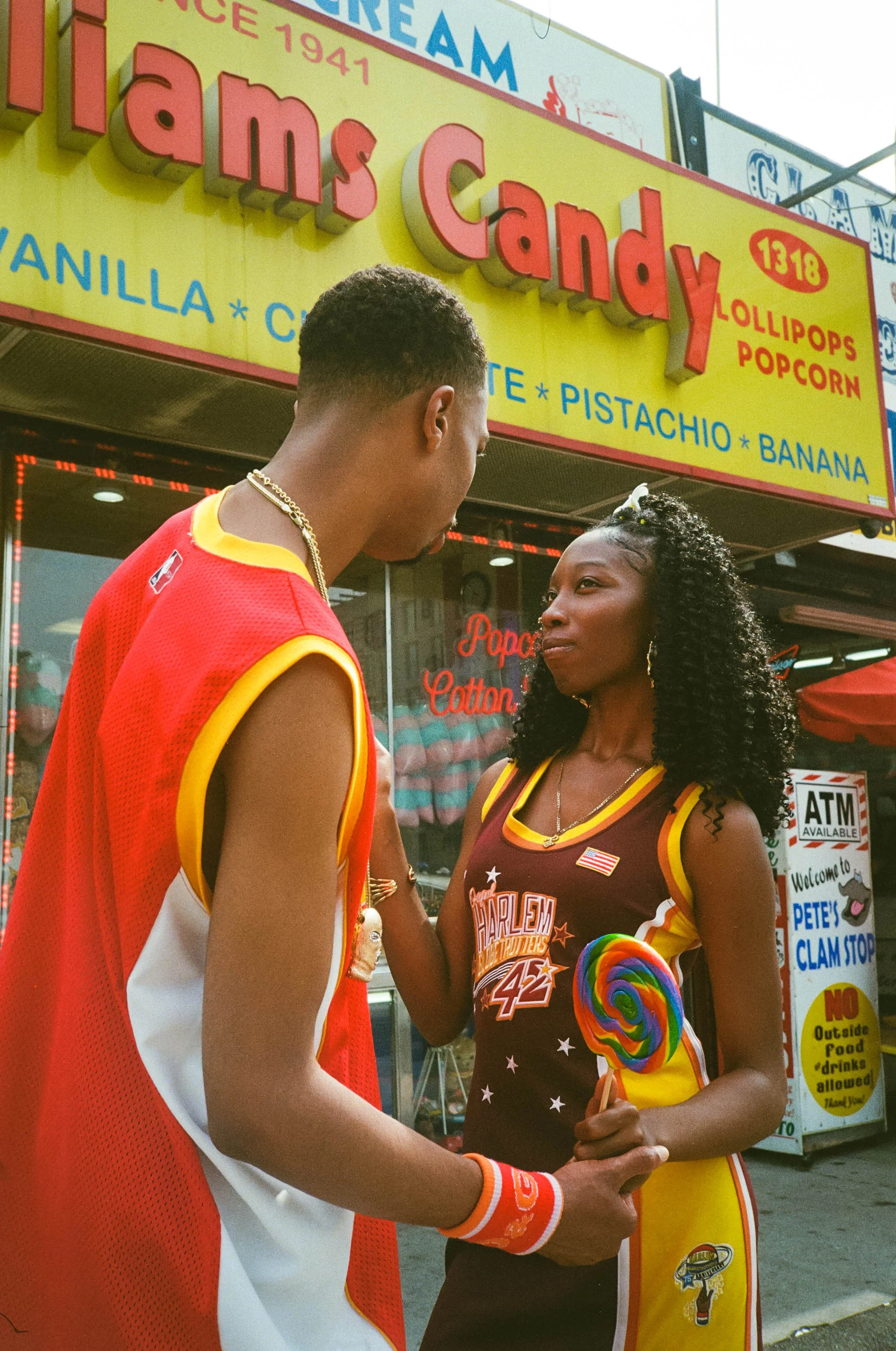 A man and a woman holding hands outside a candy shop with bright yellow and red sign. The man is wearing a basketball jersey with red, white, and yellow colors, gold chain, and wristbands. The woman is wearing a Harlem Globetrotters jersey and rainbo