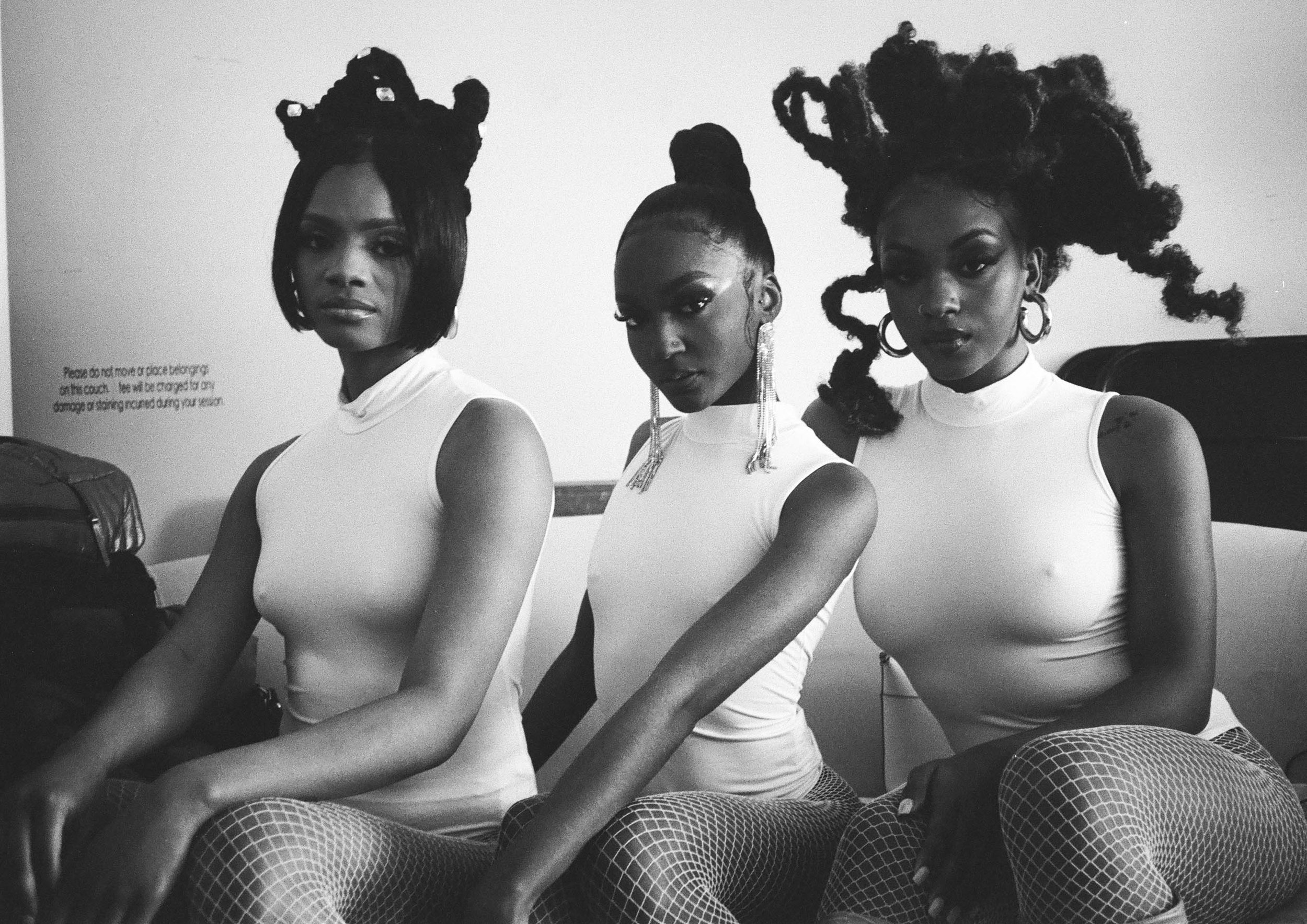 Three women with artistic hairstyles and earrings sitting in a row, wearing sleeveless tops and fishnet stockings, in a black and white photo.