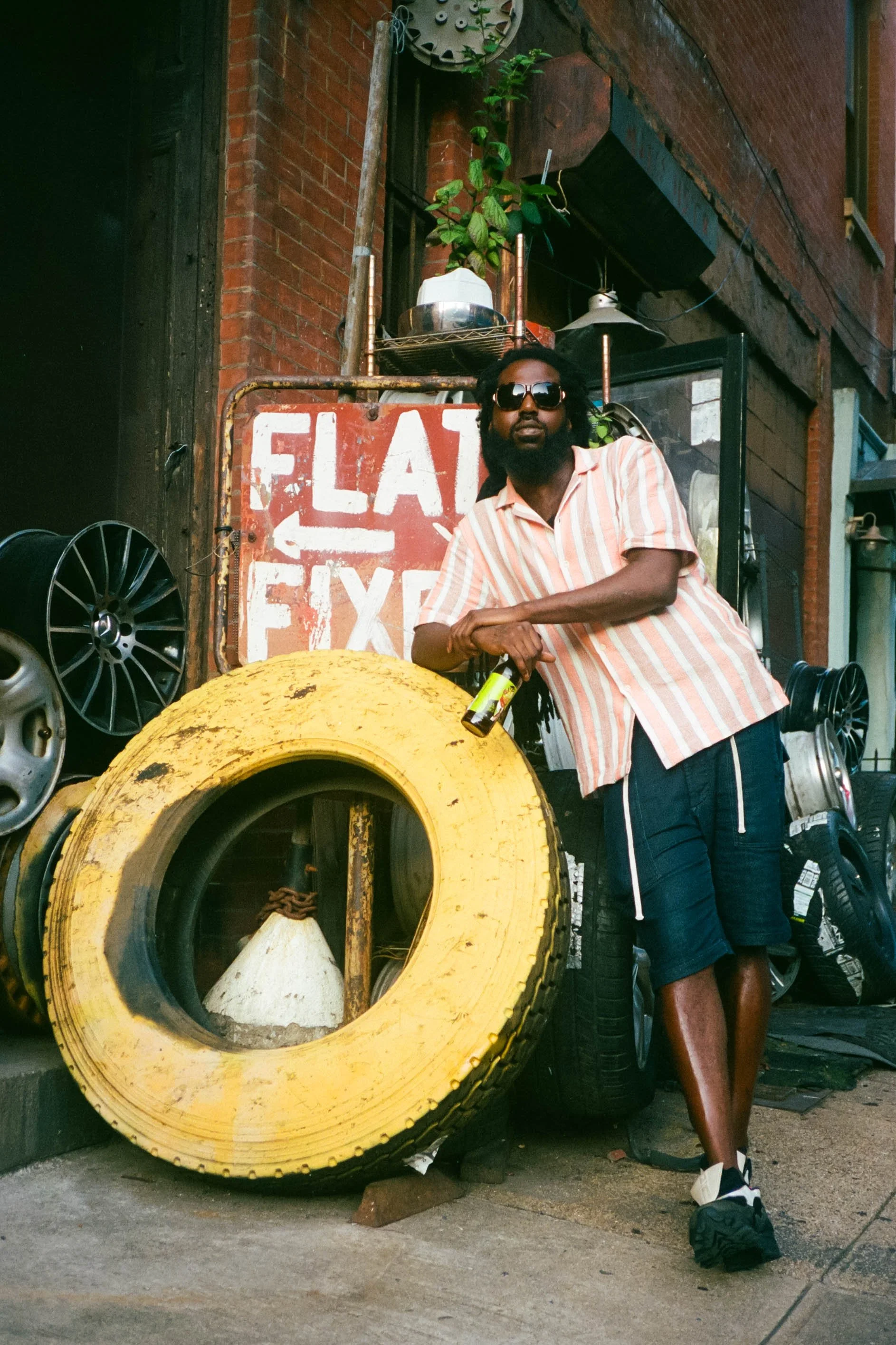 A man stands beside a large yellow tire with a white cone inside it, holding a small bottle. He wears sunglasses, a striped pink and white shirt, and dark shorts. The background features brick buildings, tires, and a sign that partially reads 'Fla...
