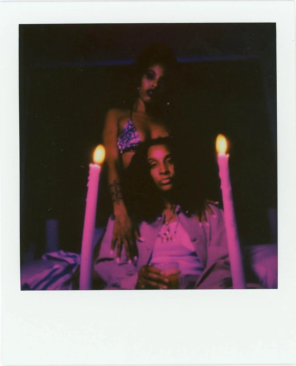 Two women in a dimly lit room with pink candles, one sitting and holding a drink, the other standing behind with dark makeup and a purple patterned top.