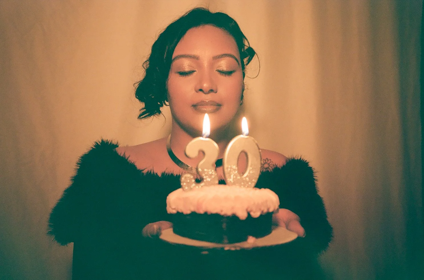 A woman with closed eyes holding a birthday cake with candles shaped as the number 20, wearing a black furry top and a choker necklace.