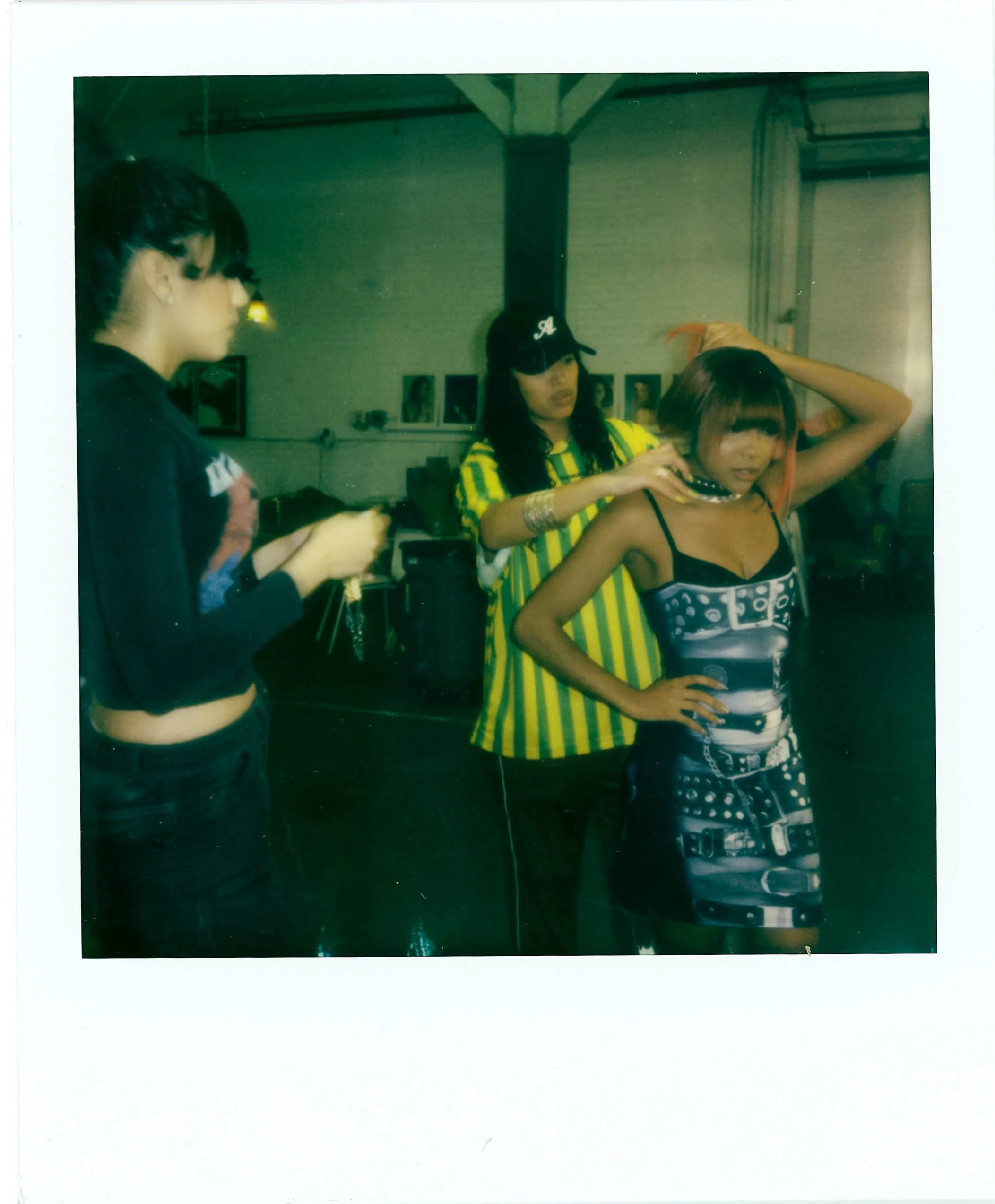 Three women in a room, with one woman applying makeup or styling another woman, who is wearing a black top with a pattern, and has her hand on her head. The room has framed pictures on the wall and hanging lights.