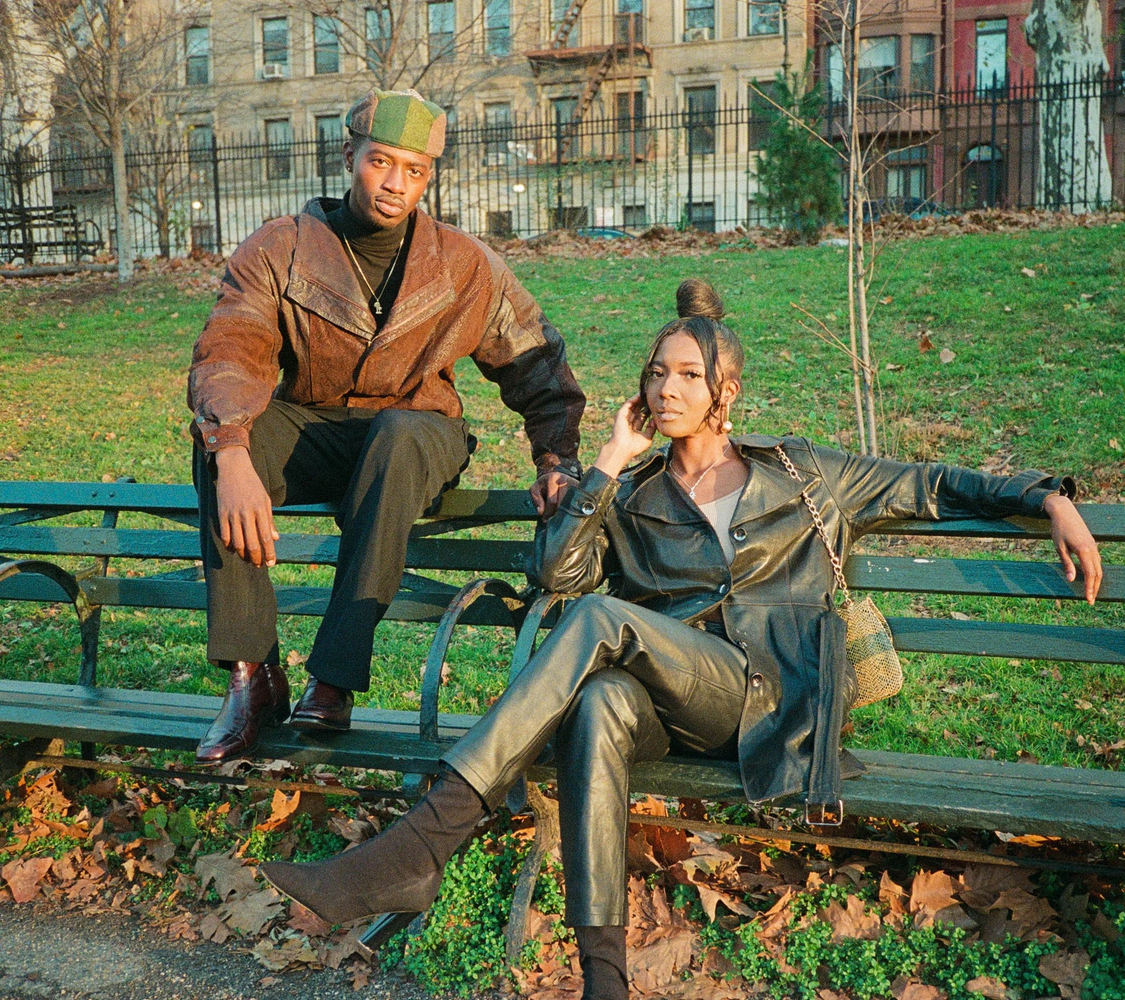 Two young adults posing on a park bench in an urban setting during fall. The man is wearing a camouflage cap, a brown leather jacket, black pants, and shiny brown boots. The woman is dressed in a black leather jacket, a white top, with a gold chain, 