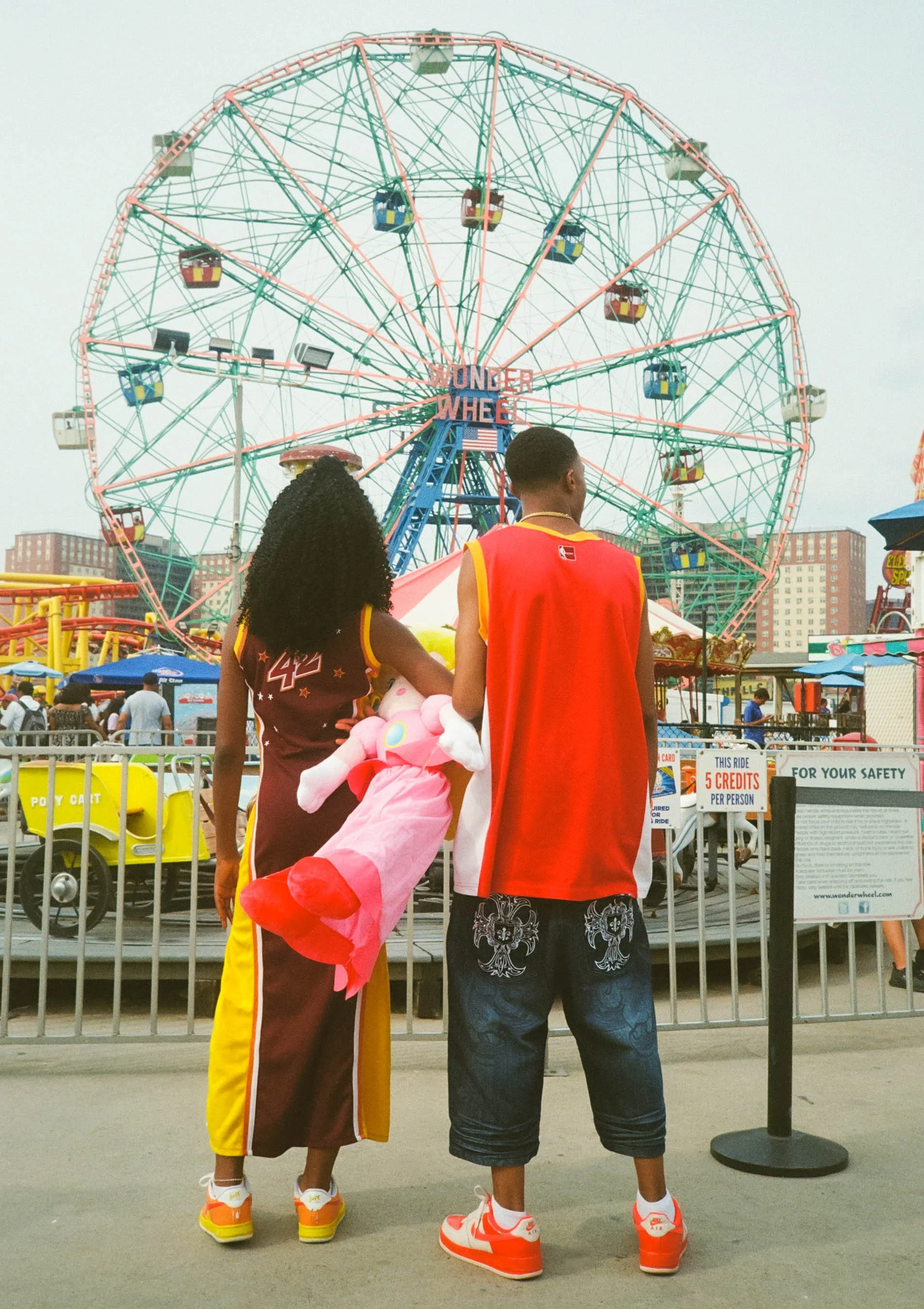 Two people wearing basketball jerseys and sneakers standing with their backs facing a Ferris wheel at an amusement park.