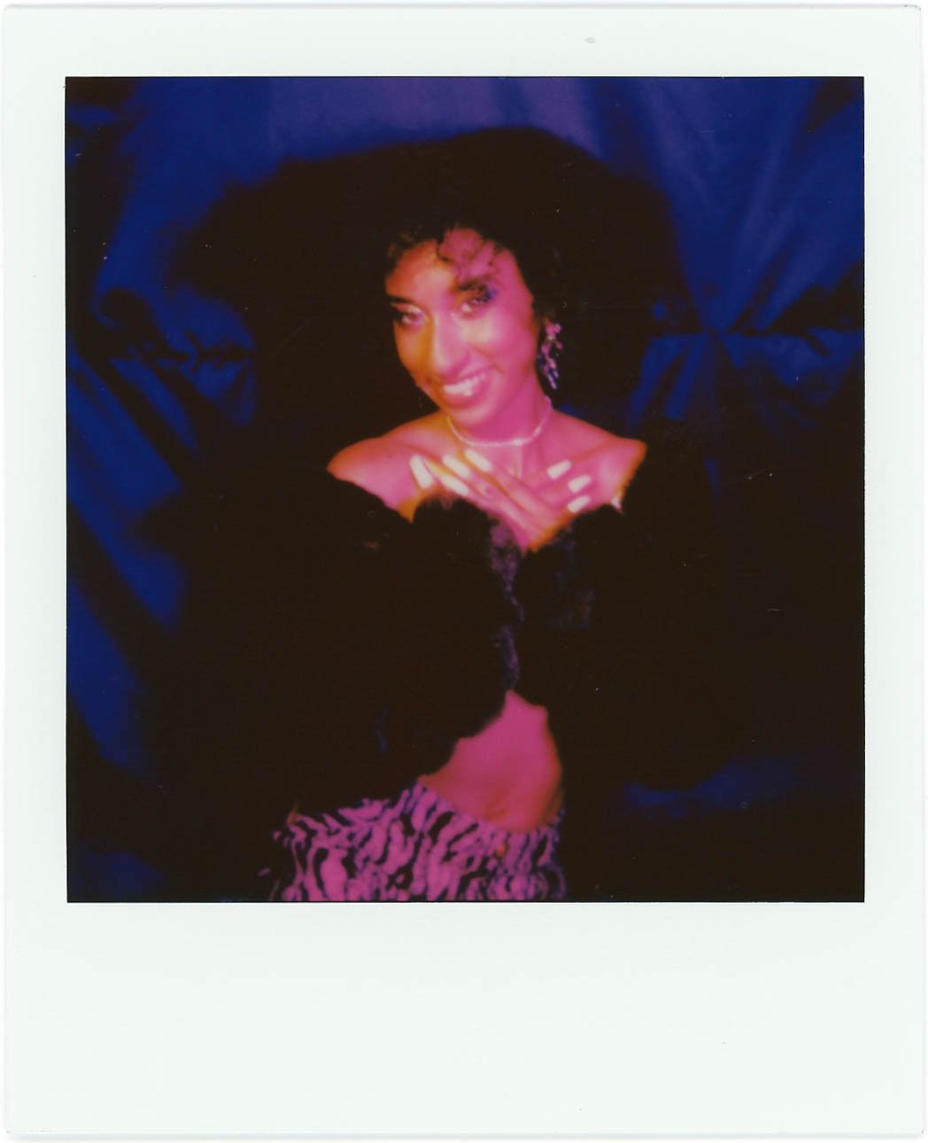 A woman with curly hair smiling, wearing jewelry and a black top, with a person in zebra print clothing partially visible in front of her, against a dark fabric background.