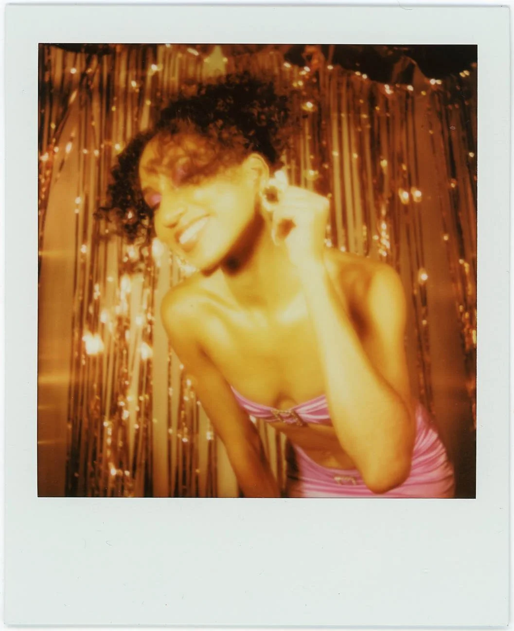A woman smiling and dancing in front of a metallic fringe curtain backdrop, wearing a pink strapless dress with a bow at the bust.
