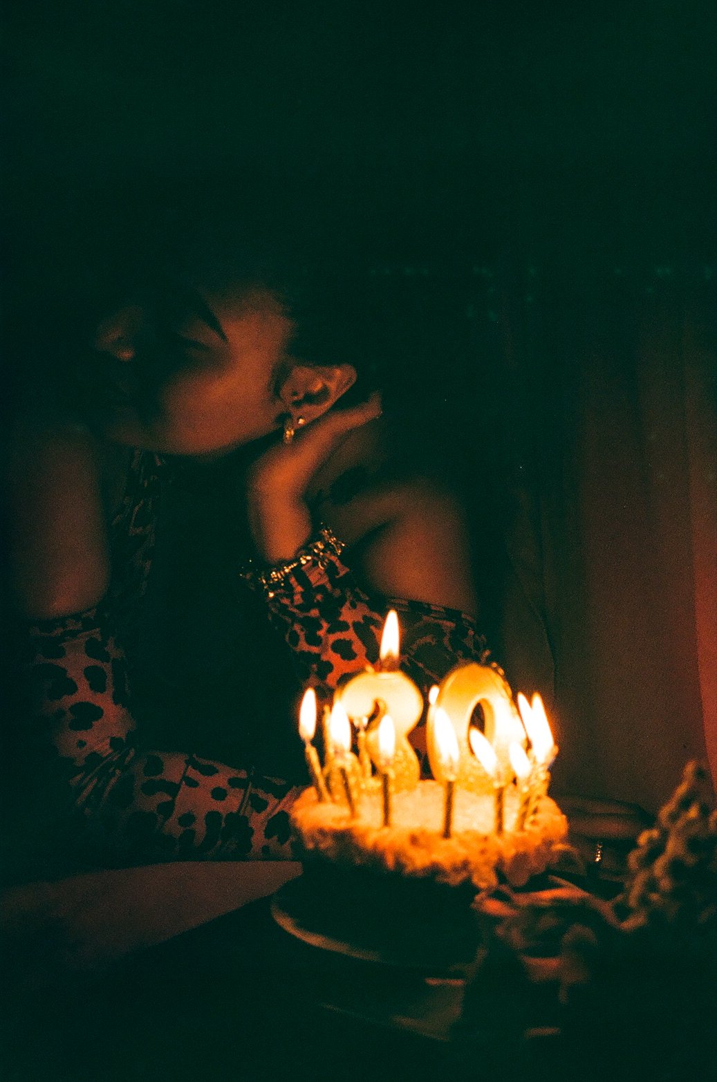 A woman celebrating her birthday with a cake featuring lit candles, with the number '30' prominently displayed. She is in a dimly lit environment, gazing at the cake, wearing jewelry and a patterned outfit.