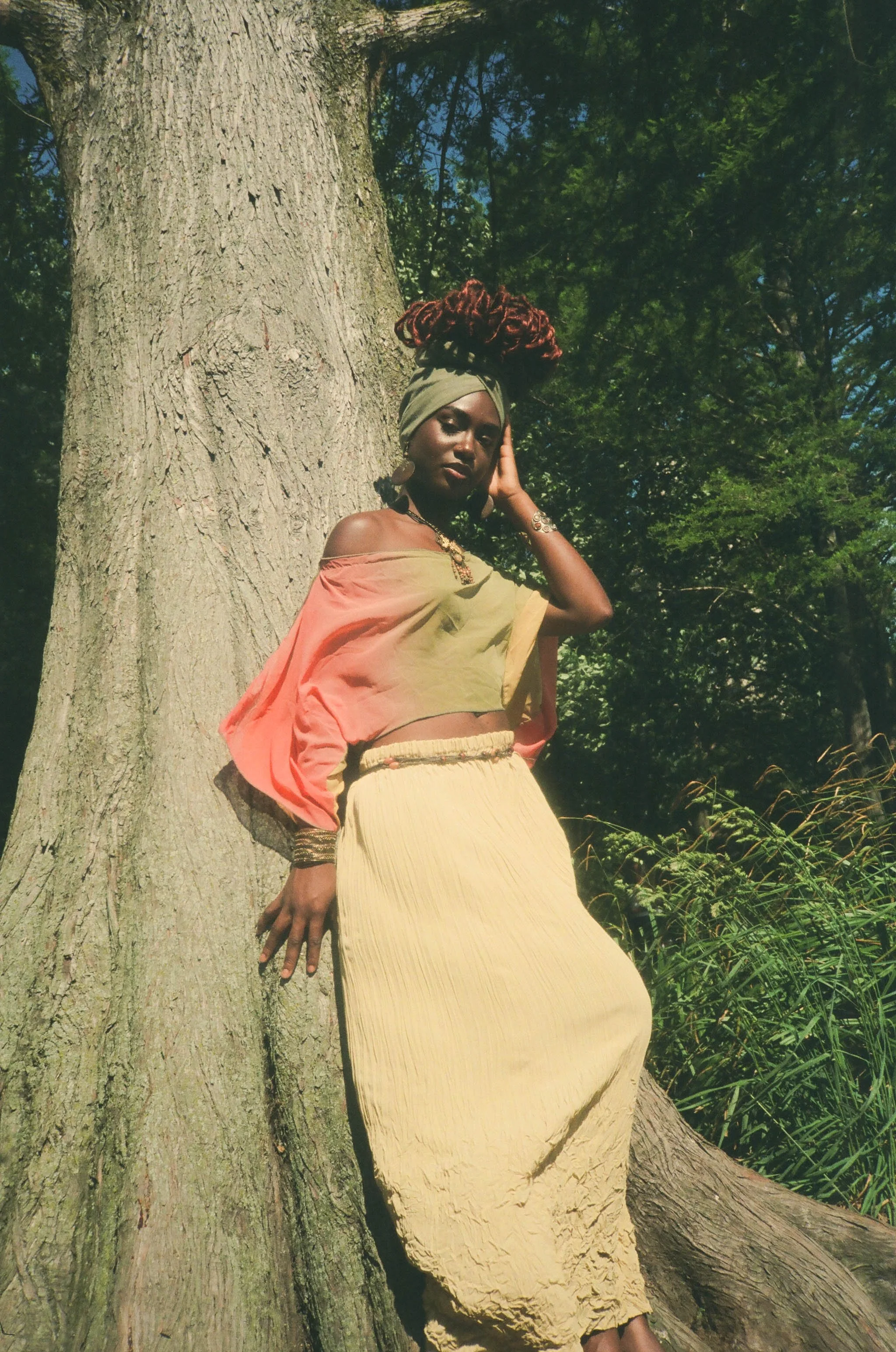 A woman with reddish dreadlocks wearing a green headwrap, earrings, and jewelry, standing outdoors beside a large tree, posing with her hand on the side of her face, dressed in a multicolored off-shoulder top and a long yellow skirt, surrounded by gr