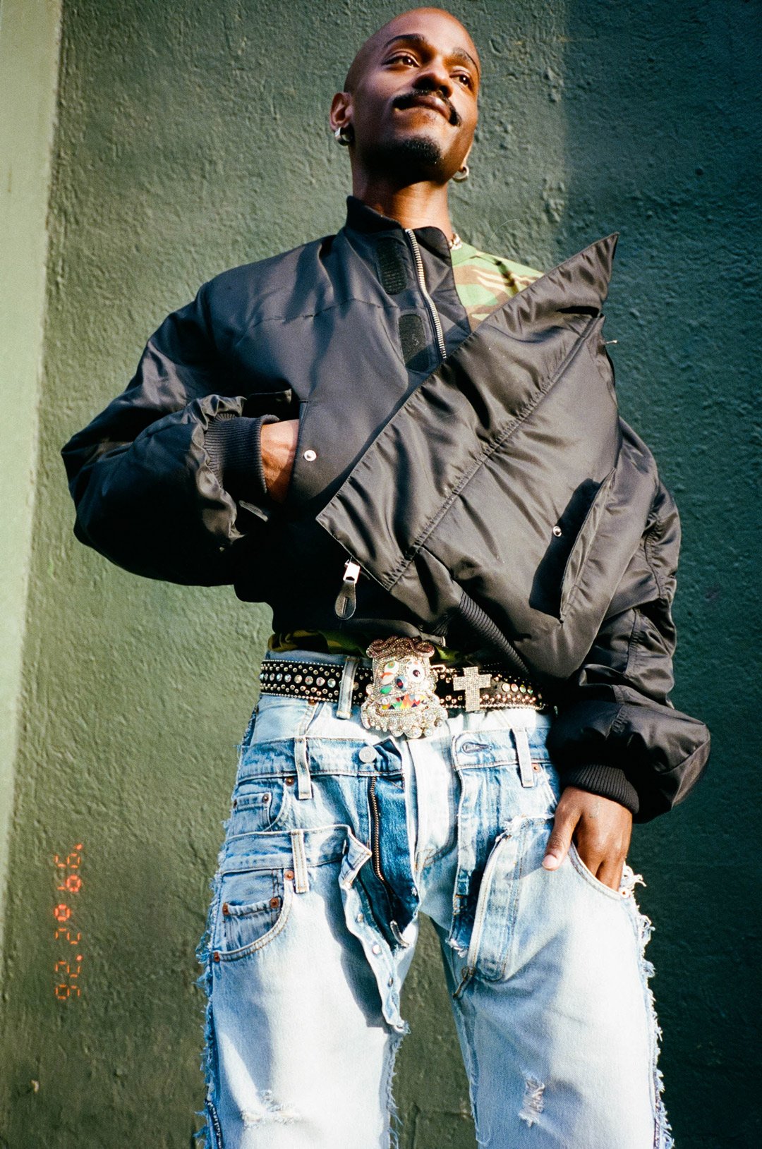 A person wearing a black bomber jacket, distressed jeans, and a belt with a decorative buckle, standing against a green wall with their hand in their pocket.