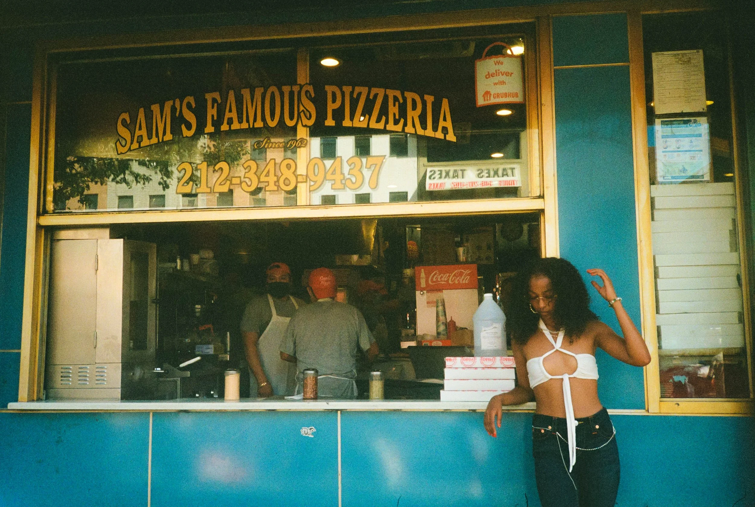 Front view of Sam's Famous Pizzeria with a large sign displaying the restaurant name and phone number, and a woman standing at the counter in the foreground.