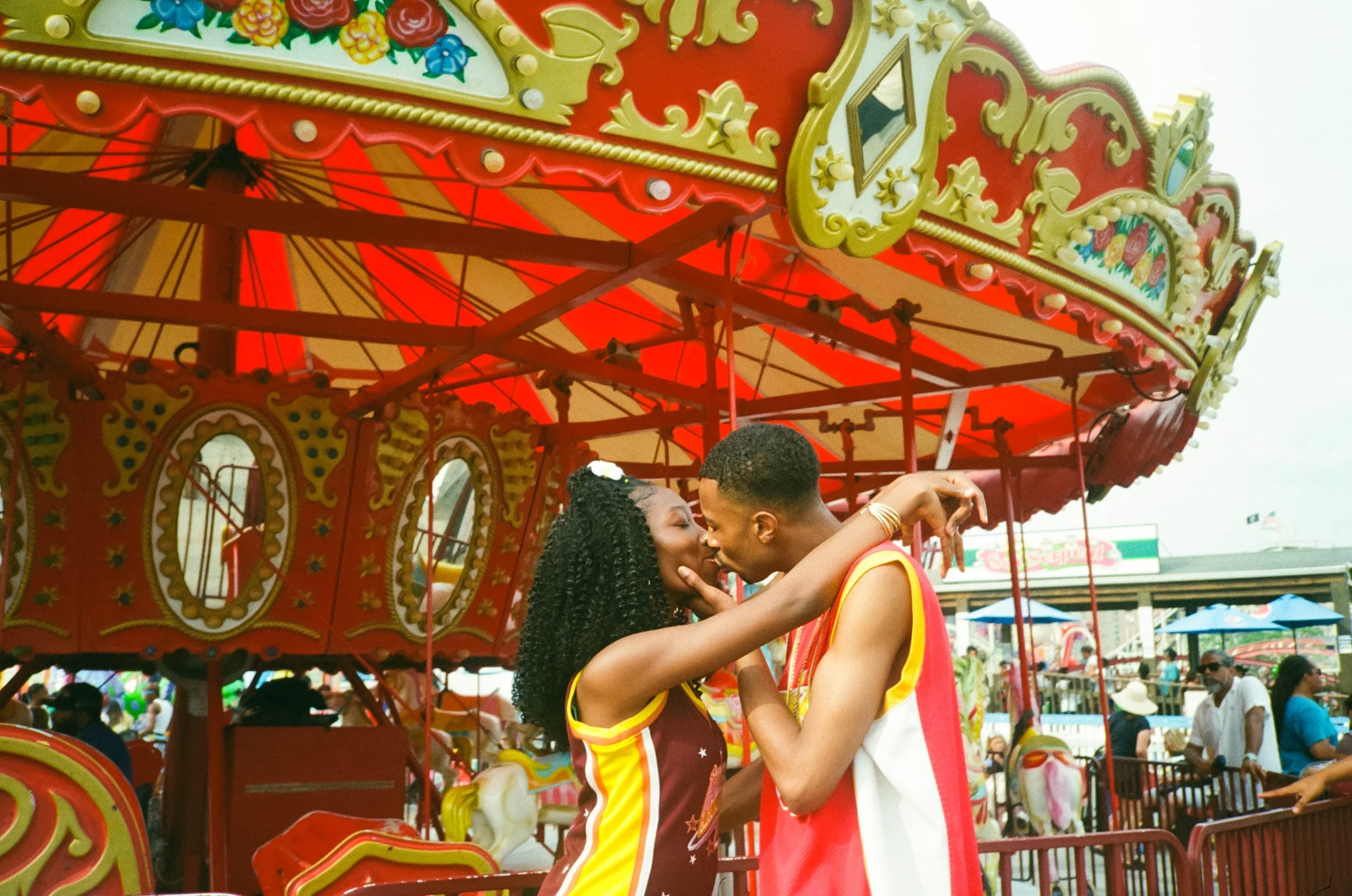 A couple kissing on a carousel ride at an amusement park, with colorful decor and a crowd in the background.
