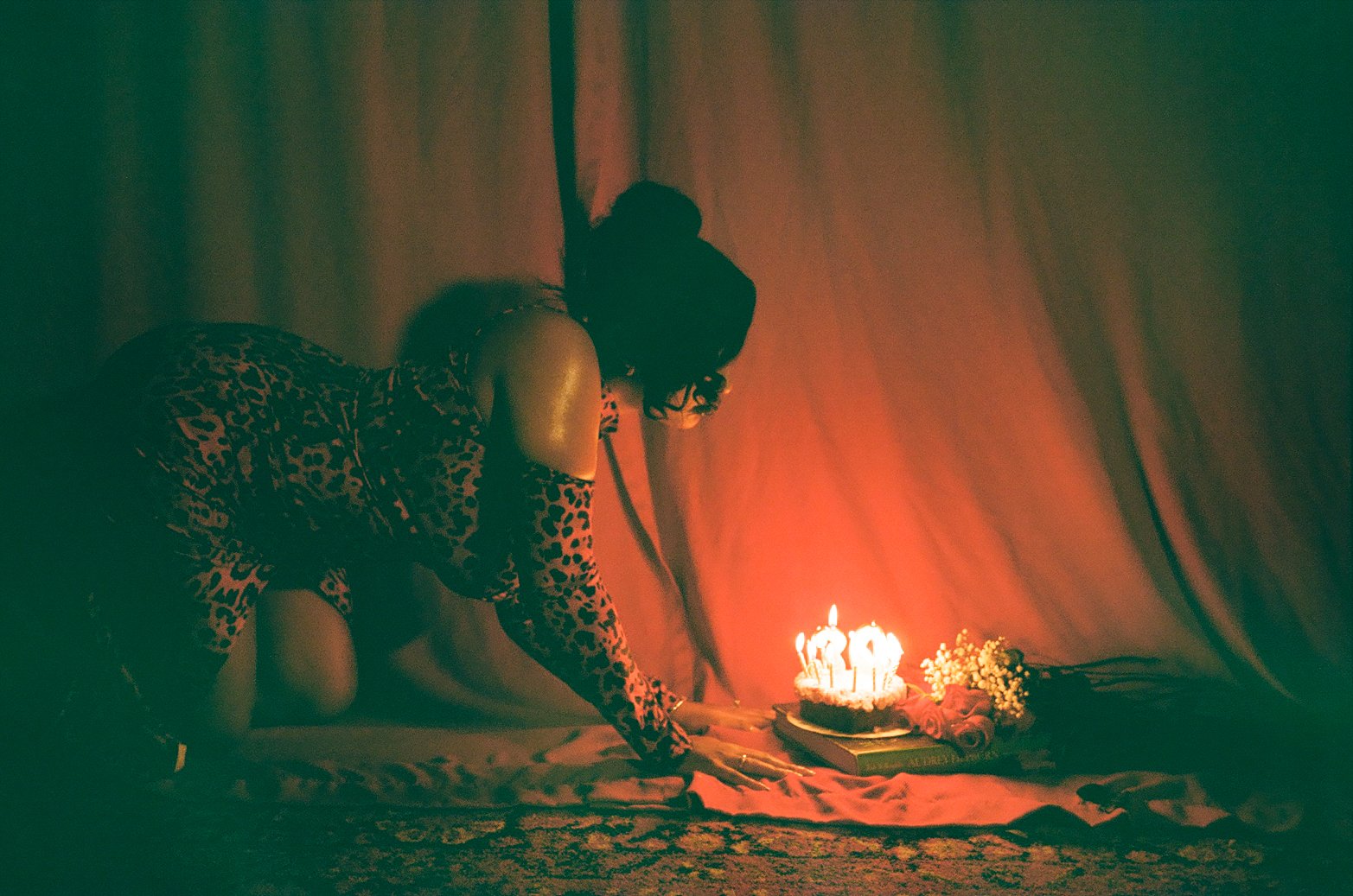 A woman wearing a leopard print dress with cut-out shoulder sleeves is kneeling on the floor, lighting birthday candles on a cake. The scene is dimly lit with the glow of the candles illuminating her and surrounding items, including red roses and a b