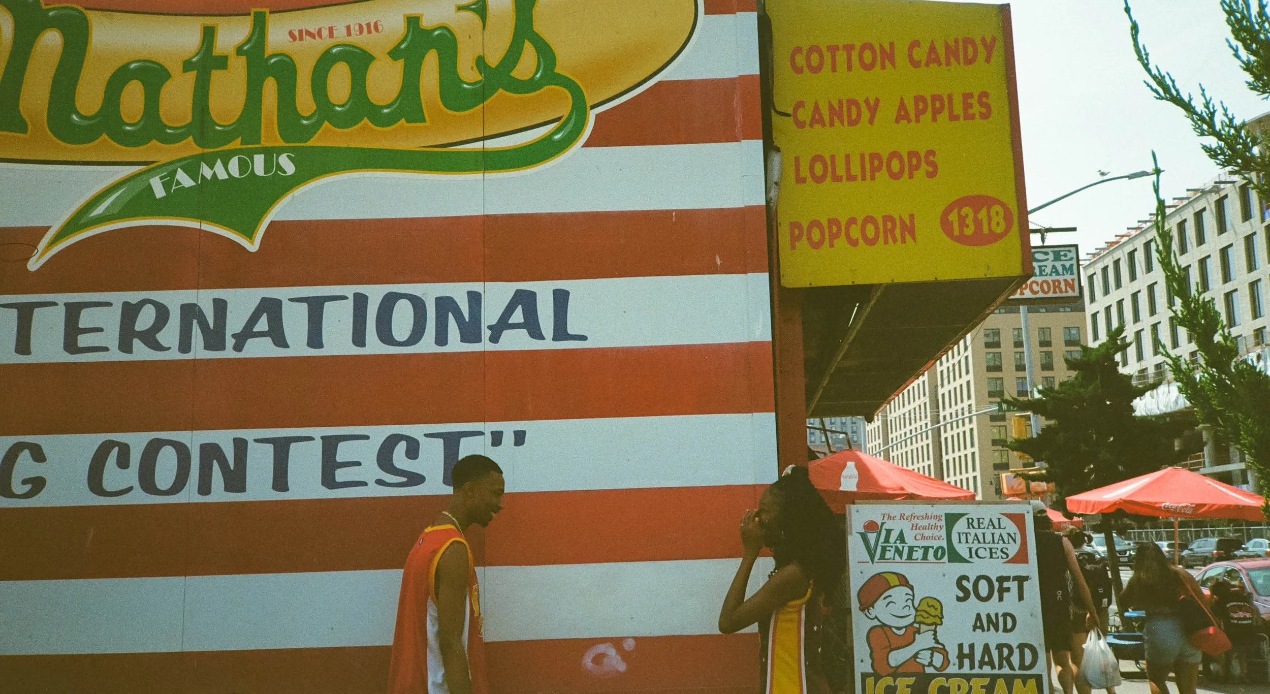 A colorful street scene featuring a large sign advertising cotton candy, candy apples, lollipops, and popcorn. There are people walking by, some wearing red and yellow outfits, with tents and building in the background.