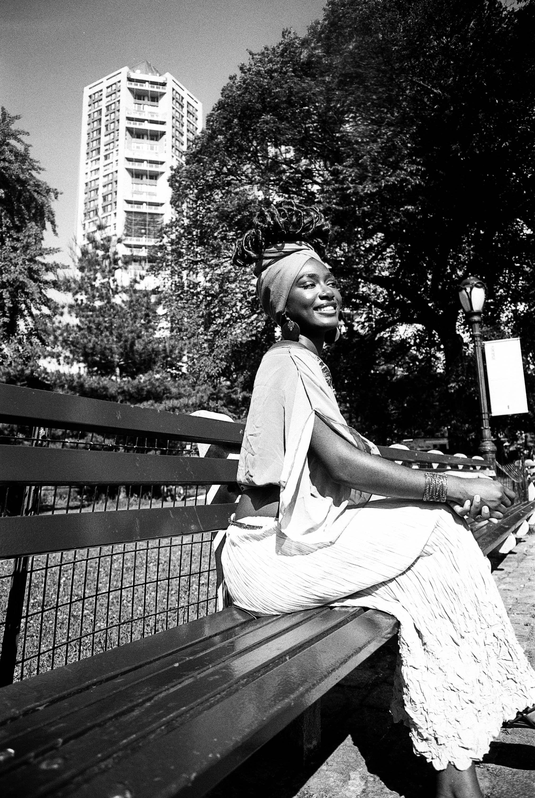 A Black woman sitting on a park bench, smiling, wearing a headwrap and earrings, with trees and a tall building in the background.