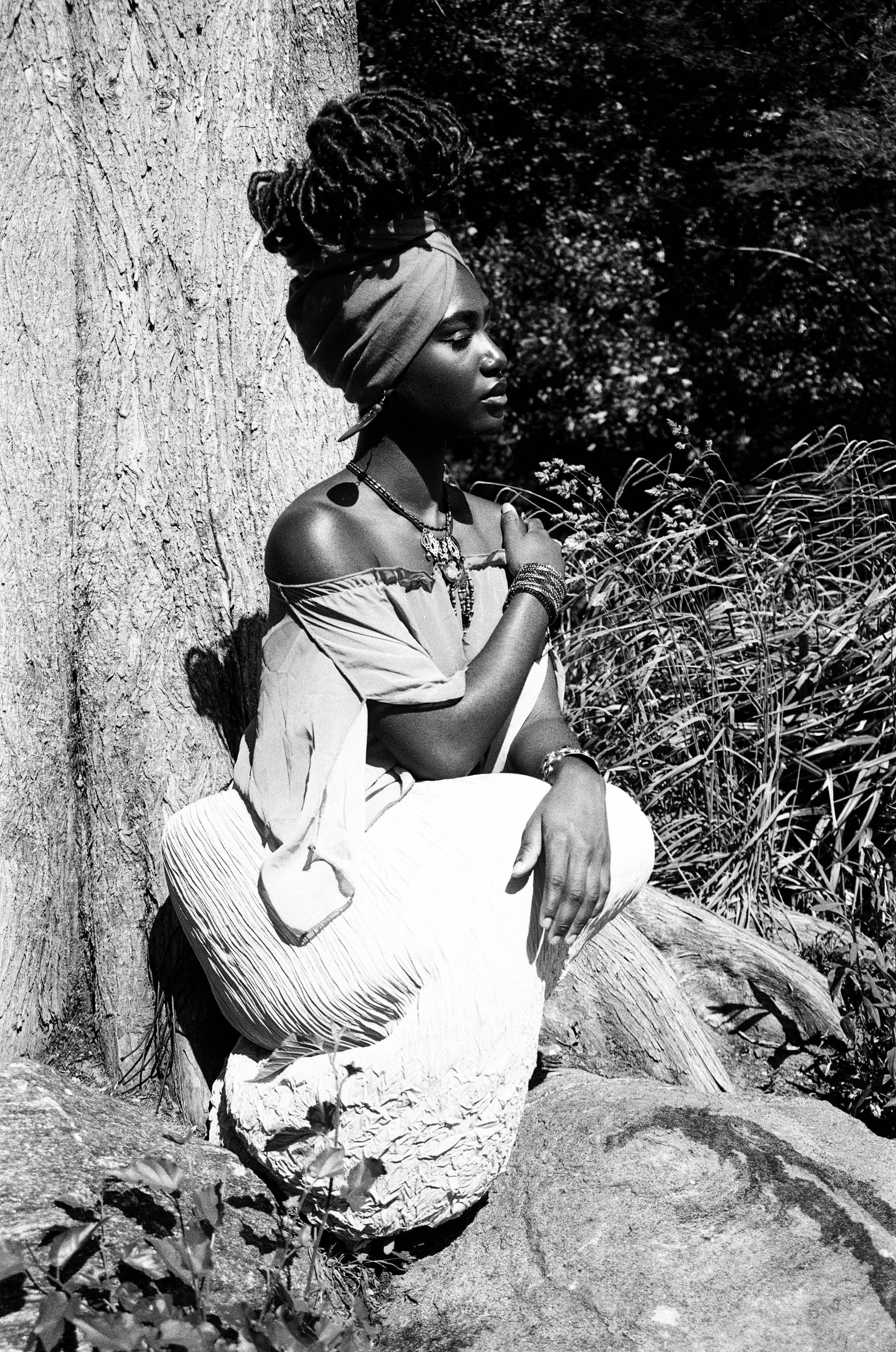 Black and white photo of a woman with dreadlocks, wearing a head wrap, off-shoulder top, and multiple necklaces, sitting against a tree with foliage around her.
