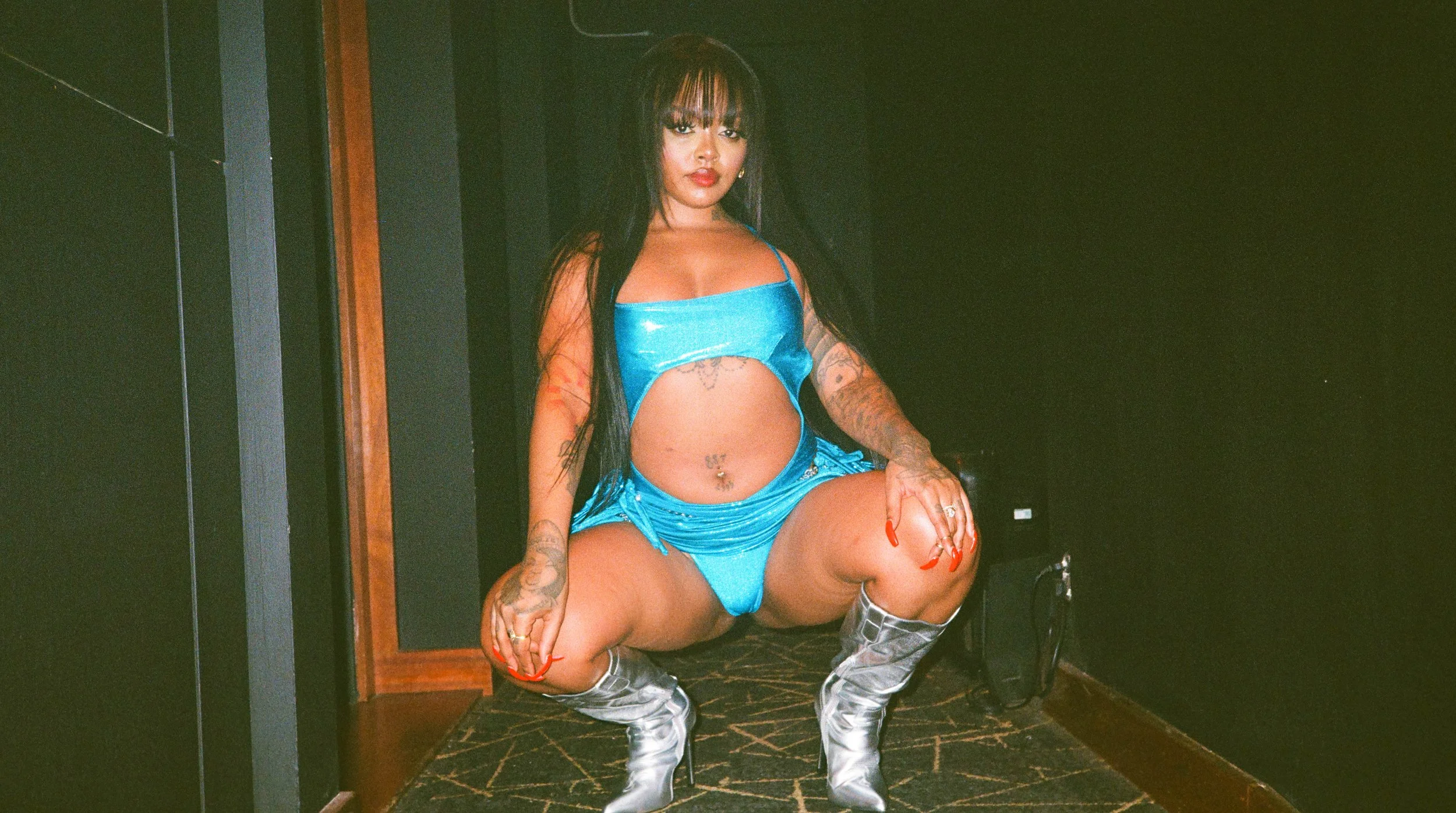 A woman dressed in a shiny blue outfit, wearing silver thigh-high boots, is seated in a squatting position with one knee raised. She has long dark hair with bangs, and visible tattoos on her arms and stomach. The background shows dark walls and a pat