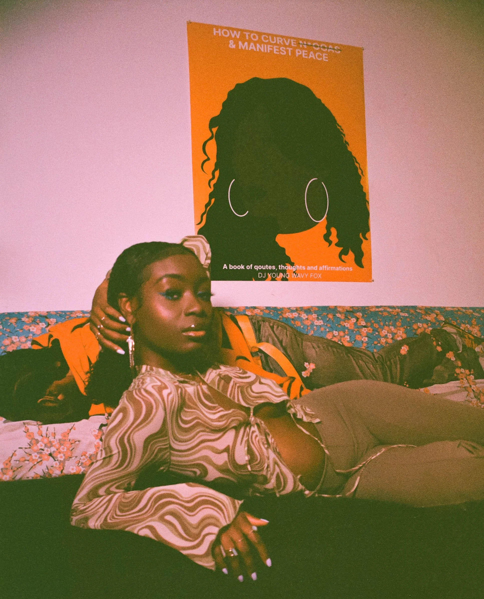 A woman lying on a floral patterned blanket in front of a colorful poster on a purple wall. The woman has dark skin, braided hair, and is wearing a zebra print jacket with an orange shirt underneath. She has jewelry and painted nails.