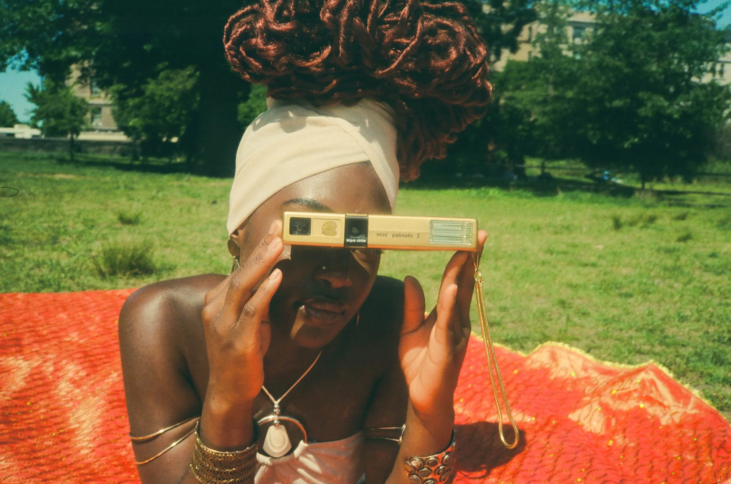 A woman with reddish curly hair, wearing a beige headband and various gold jewelry, is taking a photo using a vintage film camera outdoors on a sunny day. She is sitting on a red textured blanket in a grassy park with trees in the background.