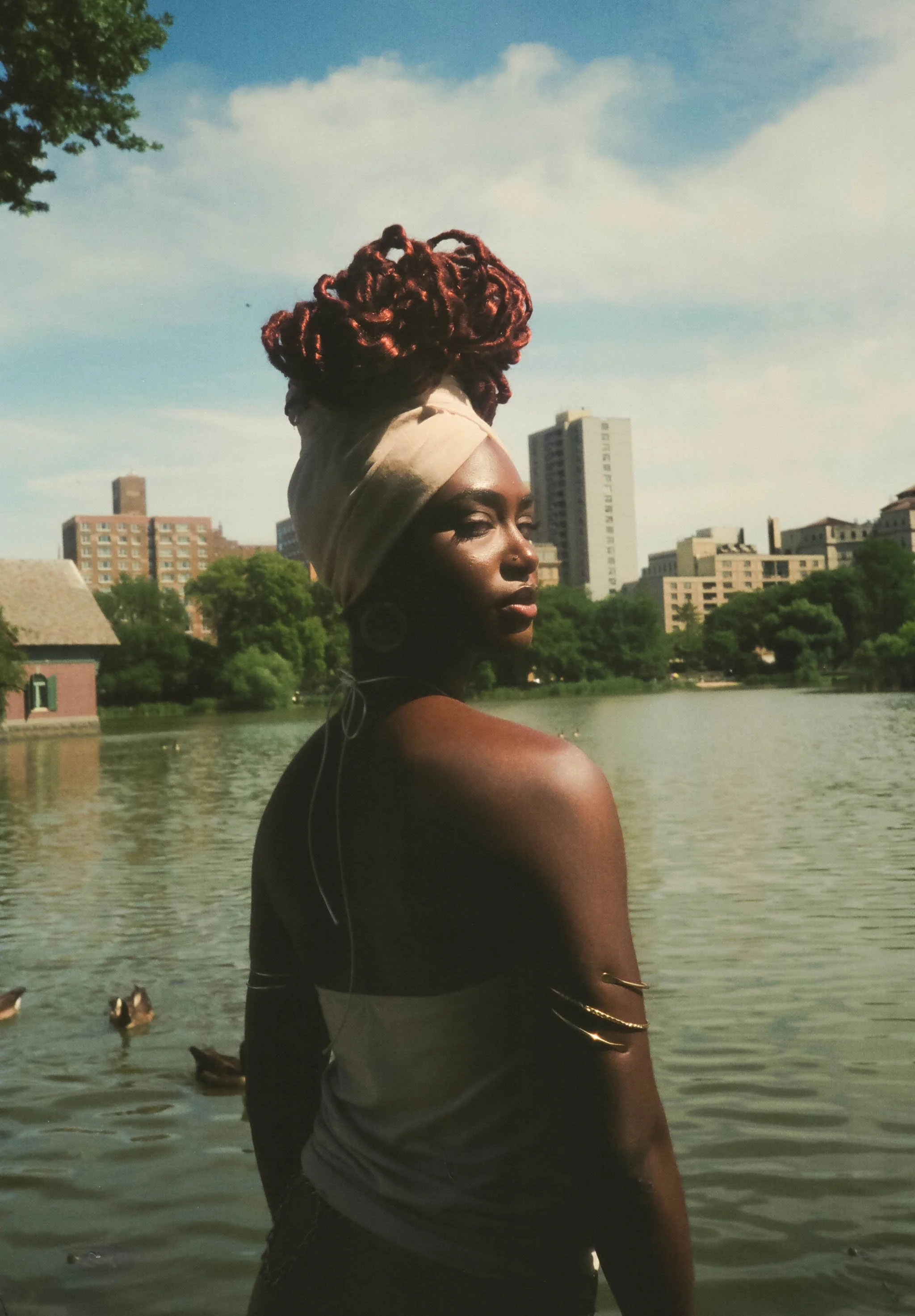 A woman with red dreadlocks with a headband standing near a river with buildings in the background and ducks in the water.