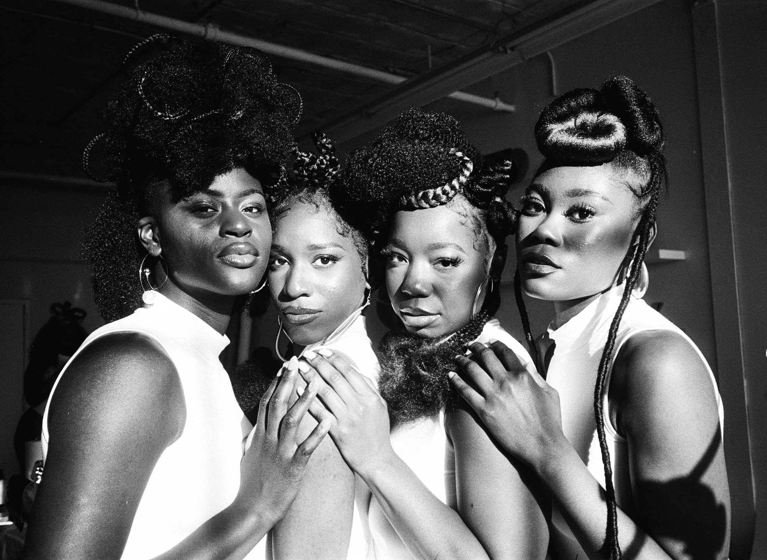 Black and white photo of five women with elaborate hairstyles, posing closely together and looking into the camera.
