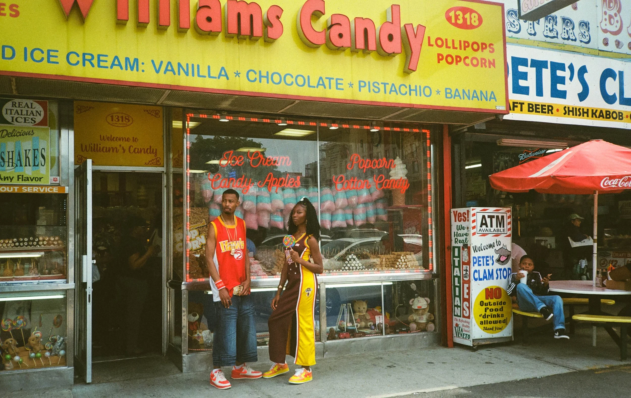 A colorful candy shop with a bright yellow sign advertising various candies and ice cream flavors. The shop has a glass window displaying cotton candy and apples, with two young people dressed in basketball jerseys standing in front. There are additi