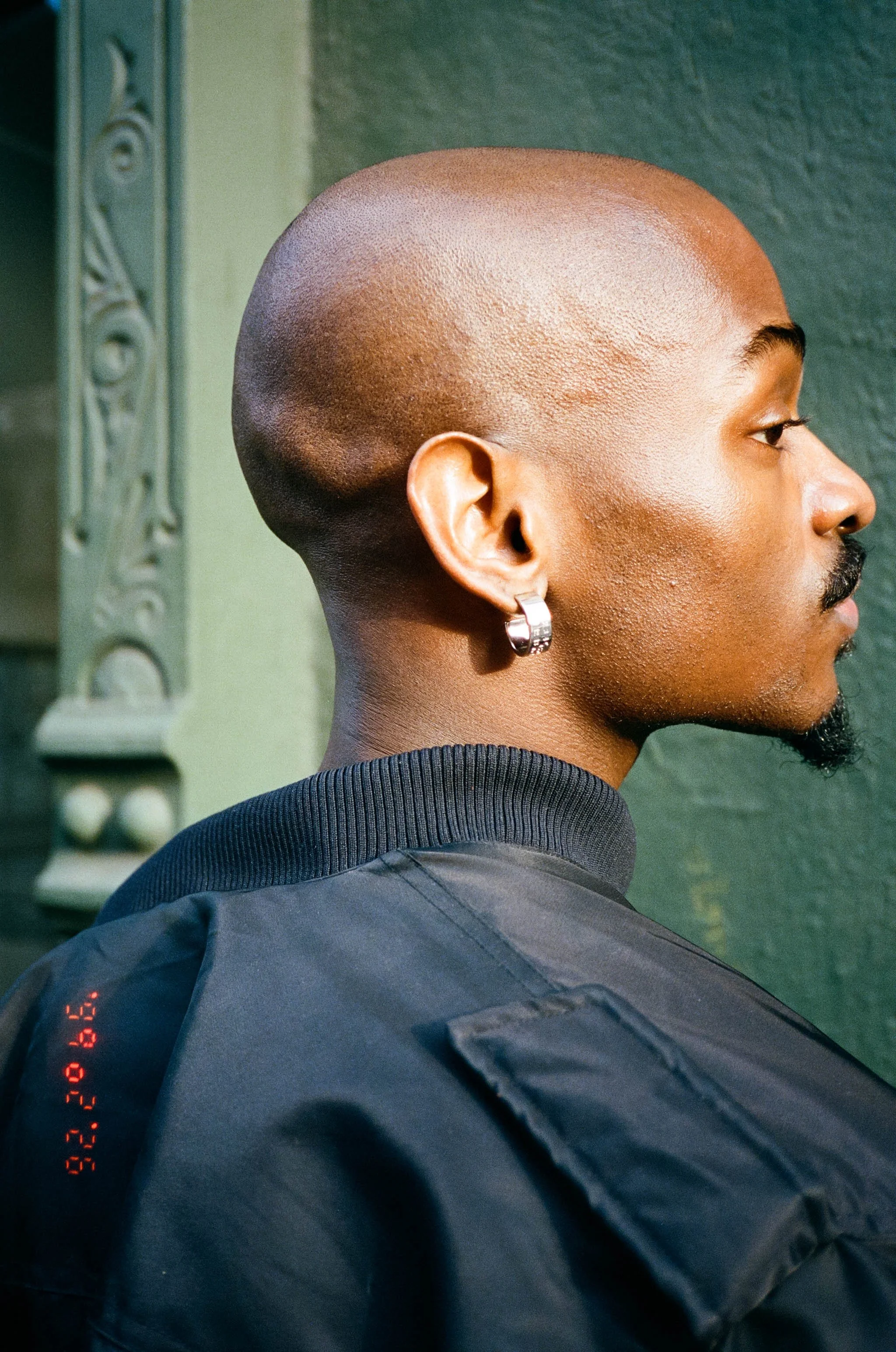 Side profile of a man with a bald head, wearing a silver hoop earring, dark jacket, and black shirt, standing in front of a green textured wall with a decorative metal element.