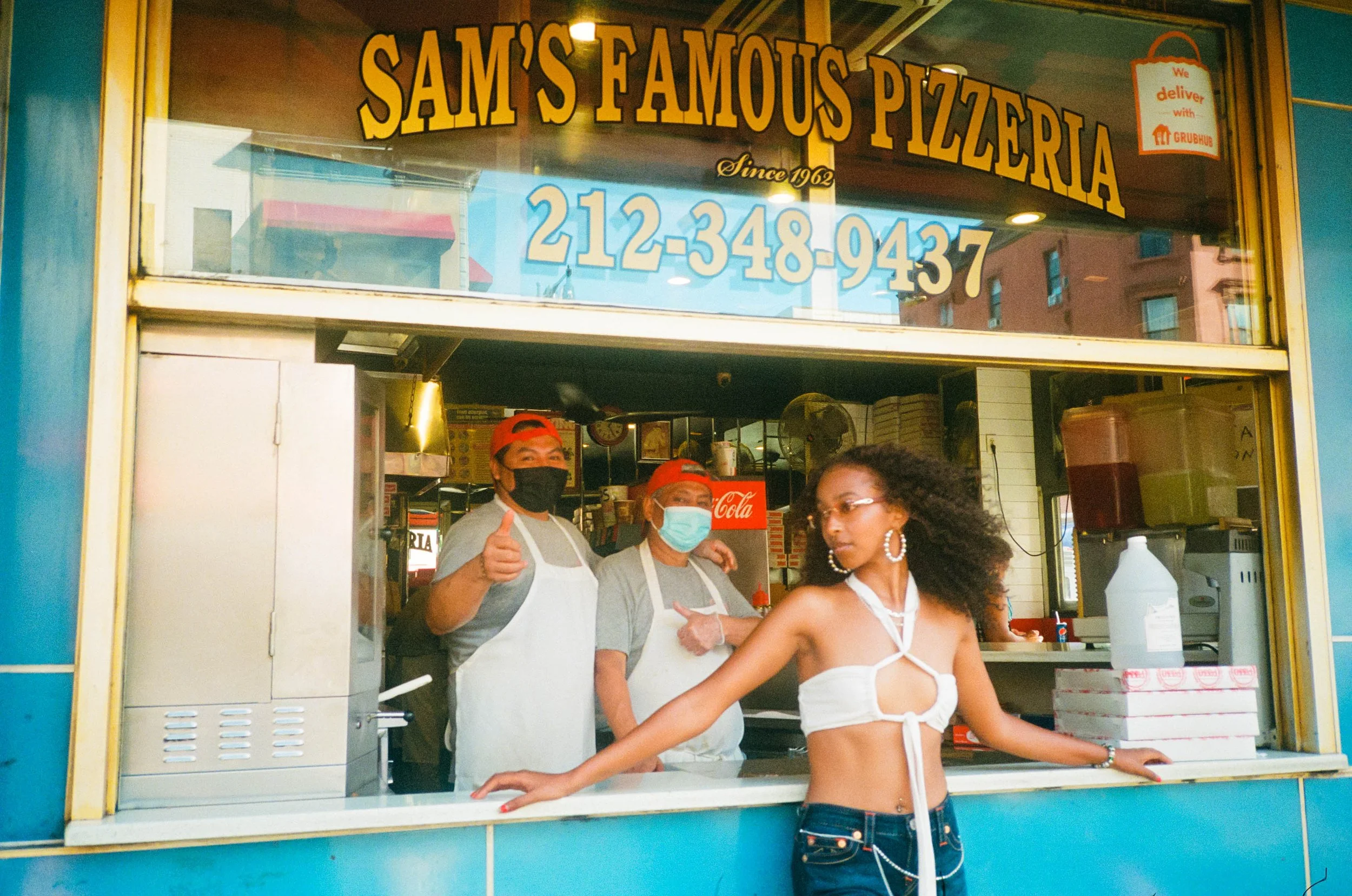 People working at a pizza stand called Sam's Famous Pizzeria, with one person giving a thumbs-up, another person wearing a face mask, and a young woman in a white top with cut-out details, standing in front of the stand.