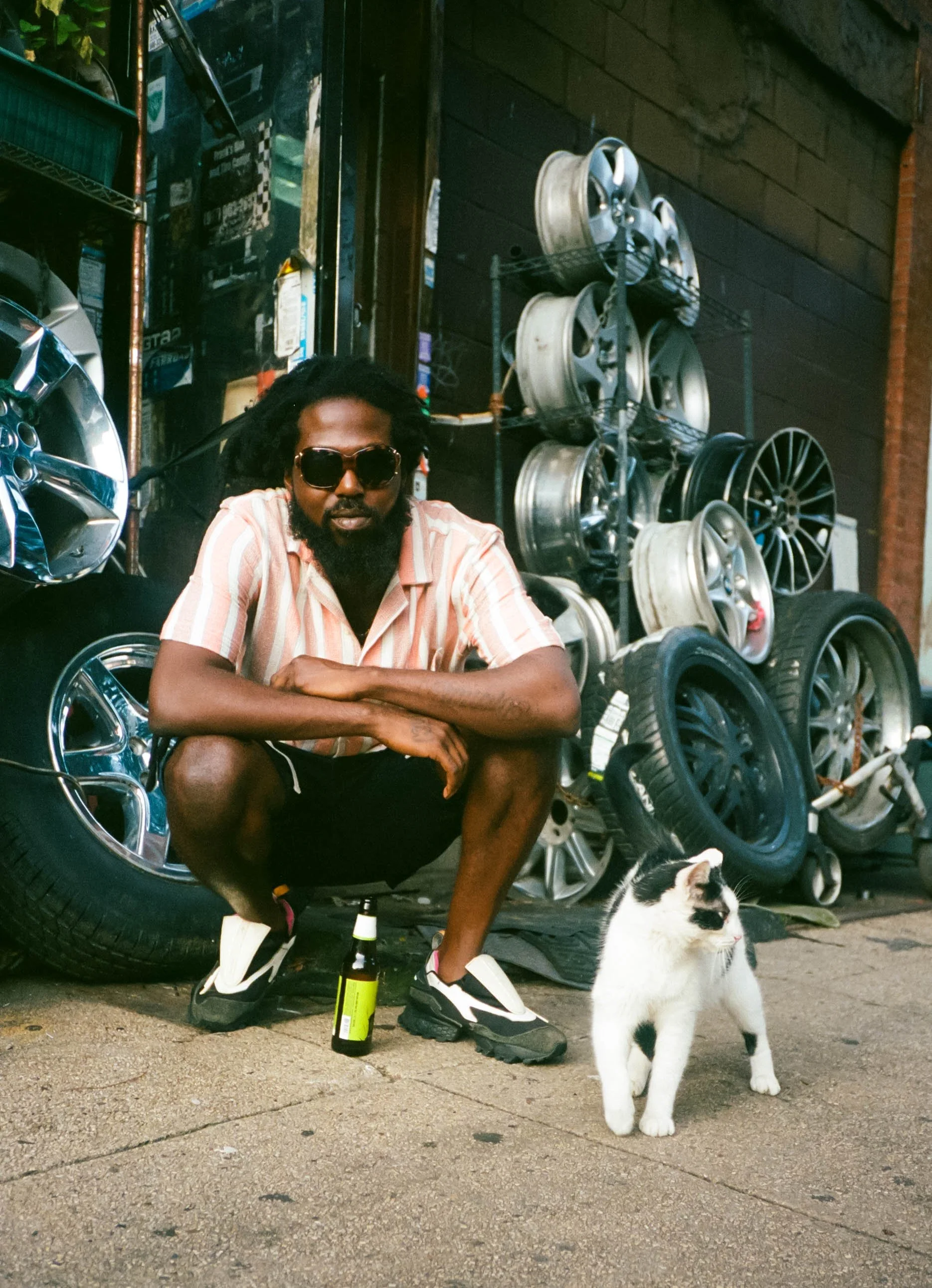 A man with dreadlocks, sunglasses, and a beard, wearing a striped shirt, black shorts, and sneakers, sitting on the ground with arms crossed. There are car rims stacked on a metal rack behind him and a cat standing on the ground nearby. Two bottles a