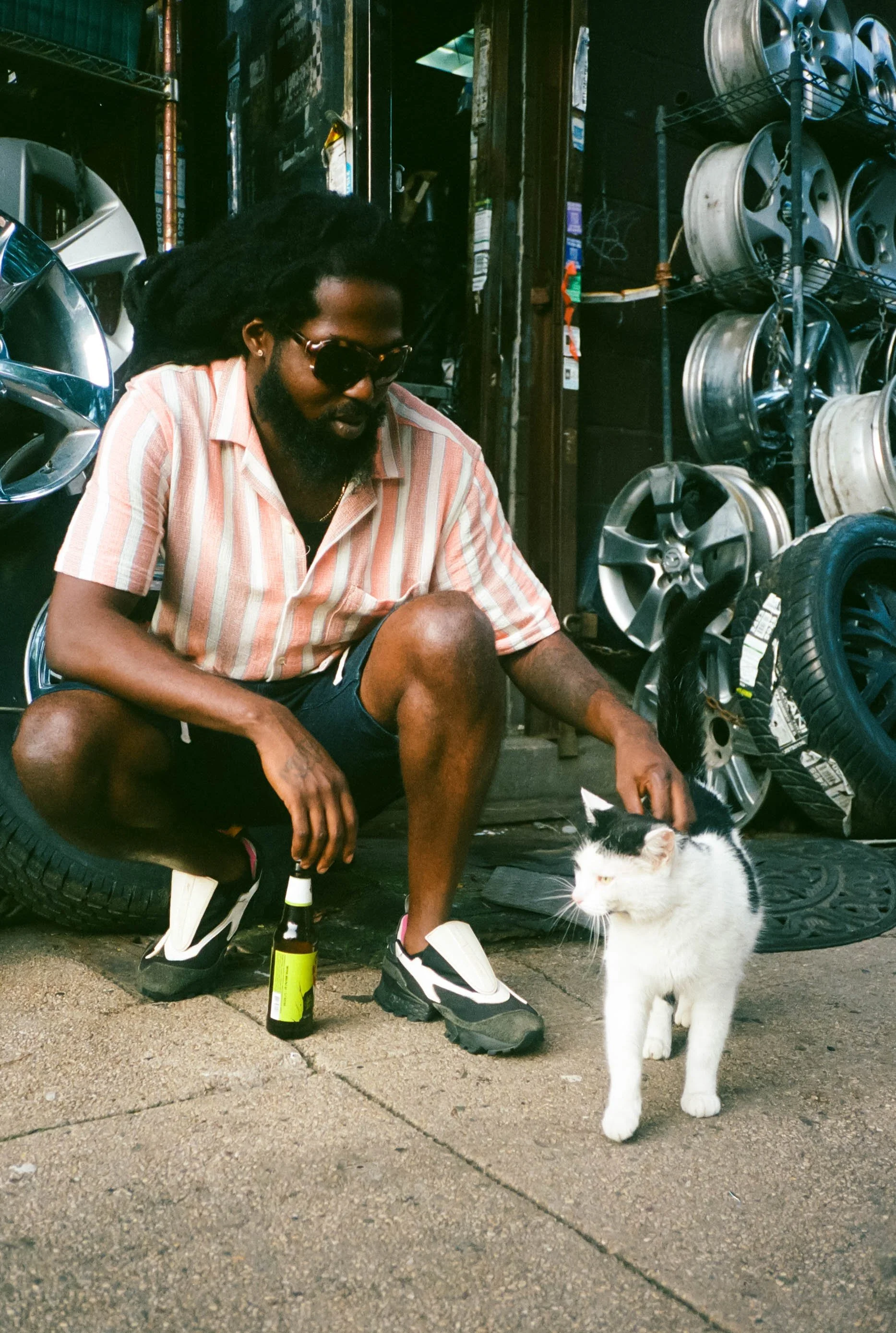 A man with dreadlocks, sunglasses, and a beard kneeling outside a tire shop next to a white and black cat, with tires stacked on shelves behind him.