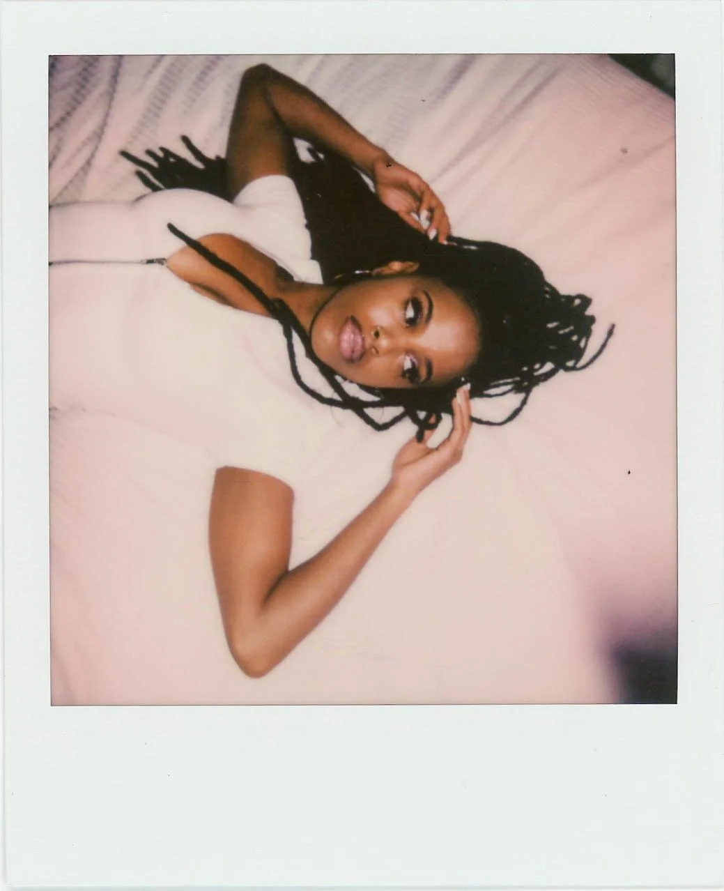 A woman with long dreadlocks lying on a bed, looking at the camera, with her hand near her head.
