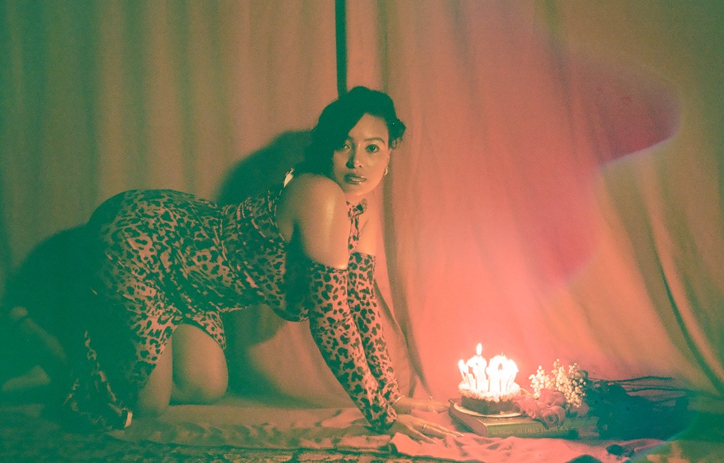 A woman in leopard print clothing and long gloves kneels on a bed facing a birthday cake with lit candles, surrounded by flowers and a book, illuminated by soft pink and orange lighting.