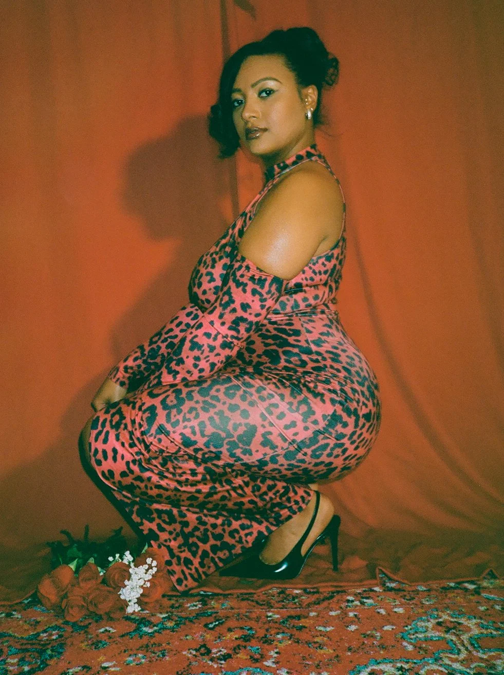 A woman in a pink and black leopard-print dress with a shoulder cutout, black high heels, earrings, and makeup, is squatting in front of an orange backdrop, with flowers on the floor.