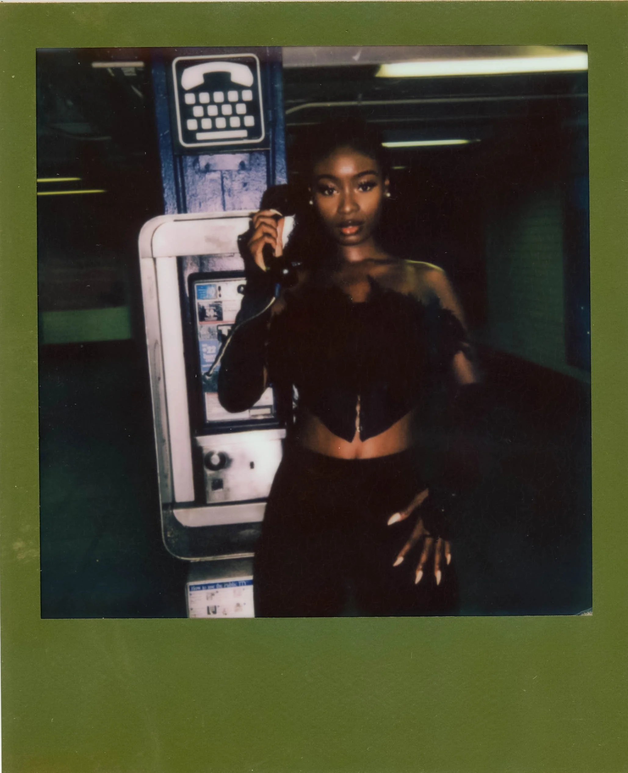 A woman with dark hair styled in a bun and wearing dark clothing is using a payphone on a green wall at night. There is an ATM or vending machine in the background.
