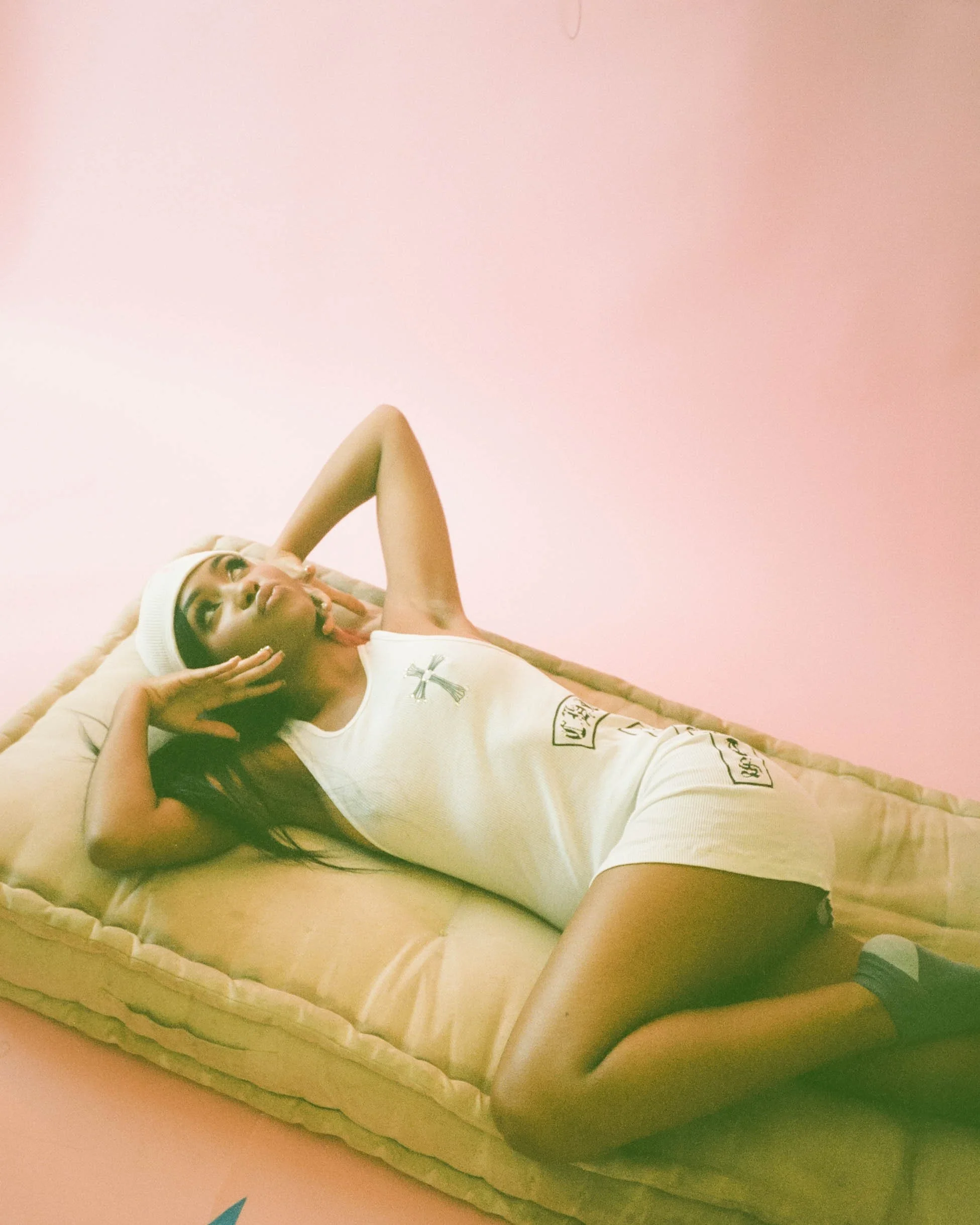 A young woman lying on a bed with a pillow, wearing a white tank top and a white beanie, resting her head on her hand, on a bedroom with pink walls.