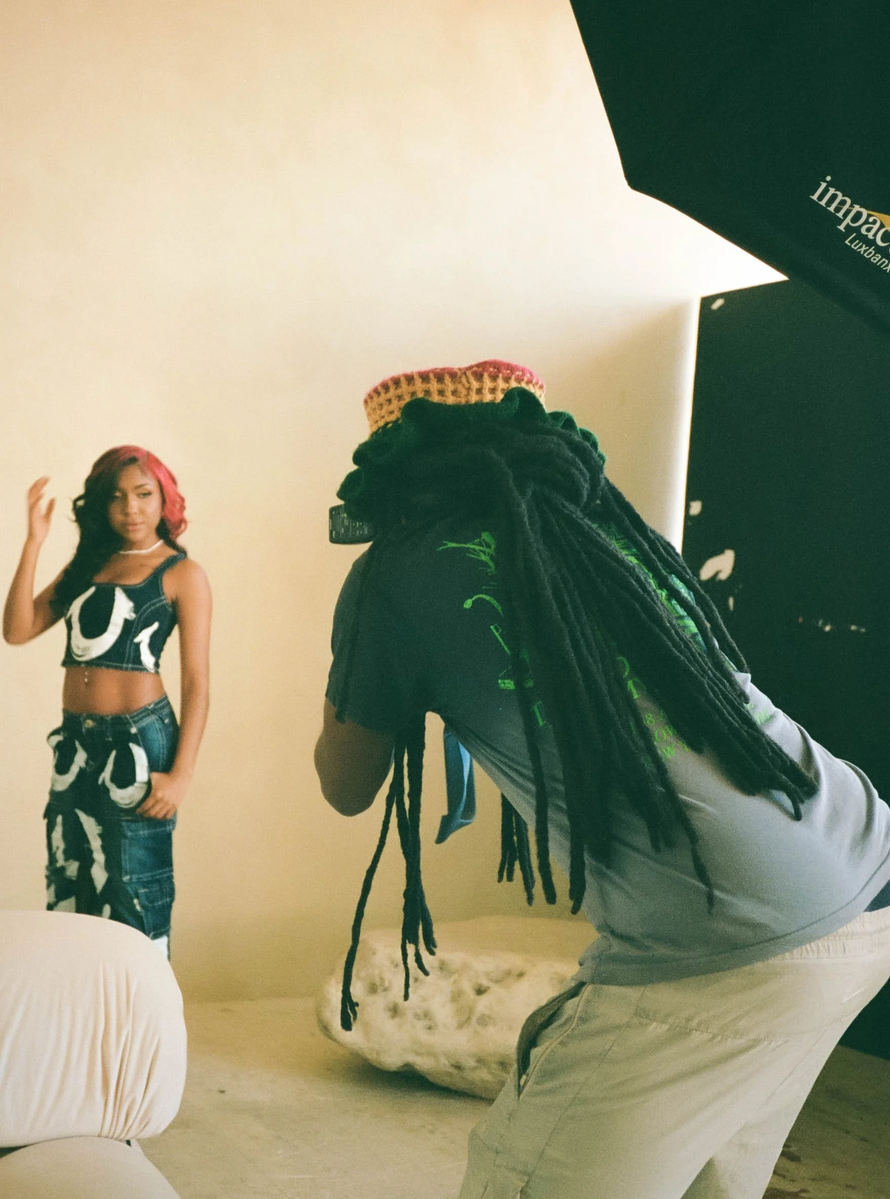 A photographer with long dreadlocks wearing a gray jacket is taking a photo of a woman with red hair in a studio. The woman is wearing a black and white striped crop top and jeans, standing with her hand on her hip, posing for the camera. The studio 