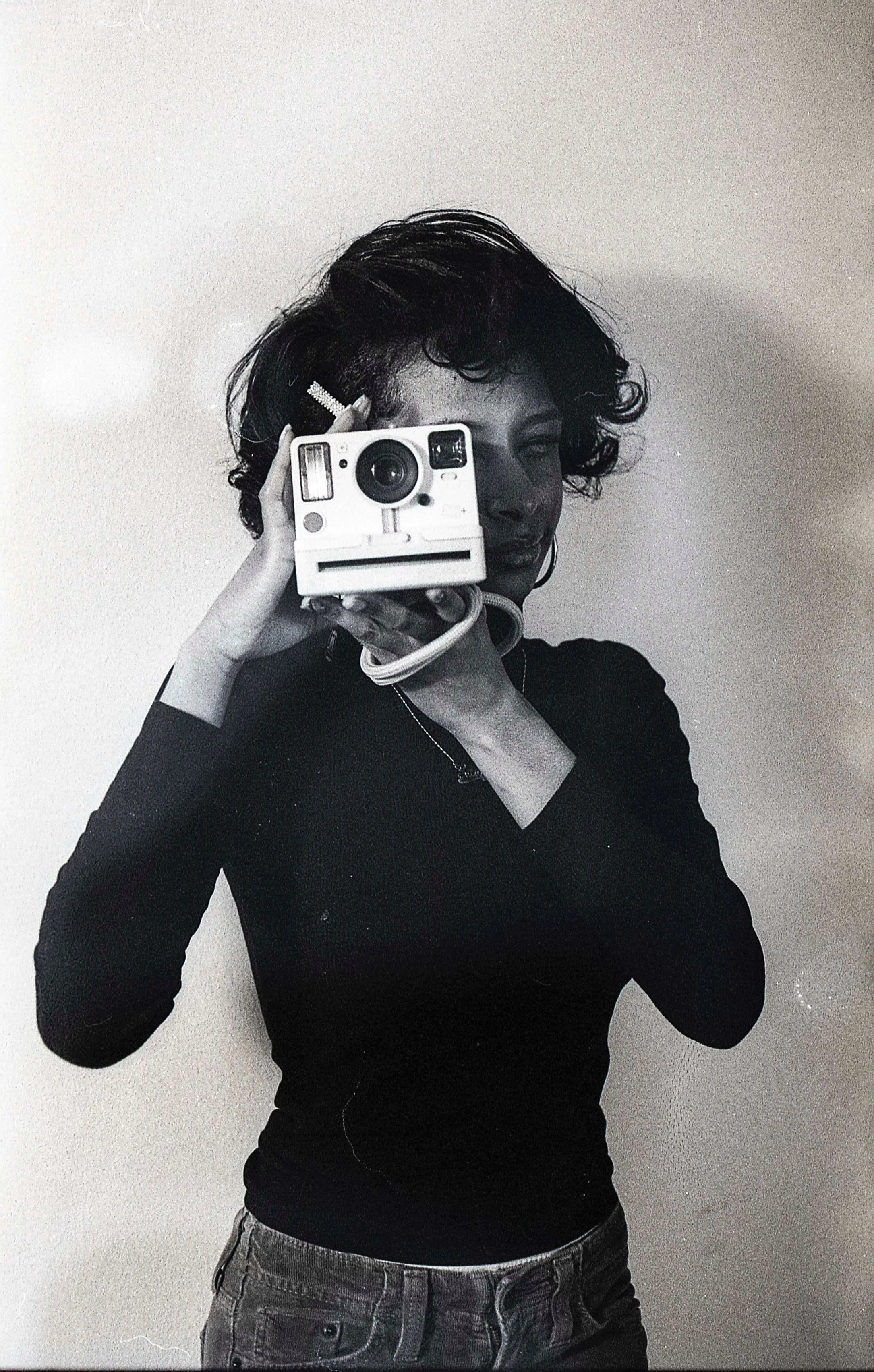Black and white photo of a woman taking a mirror selfie with a Polaroid camera. She has short curly hair, is wearing a dark long-sleeve top and denim shorts.