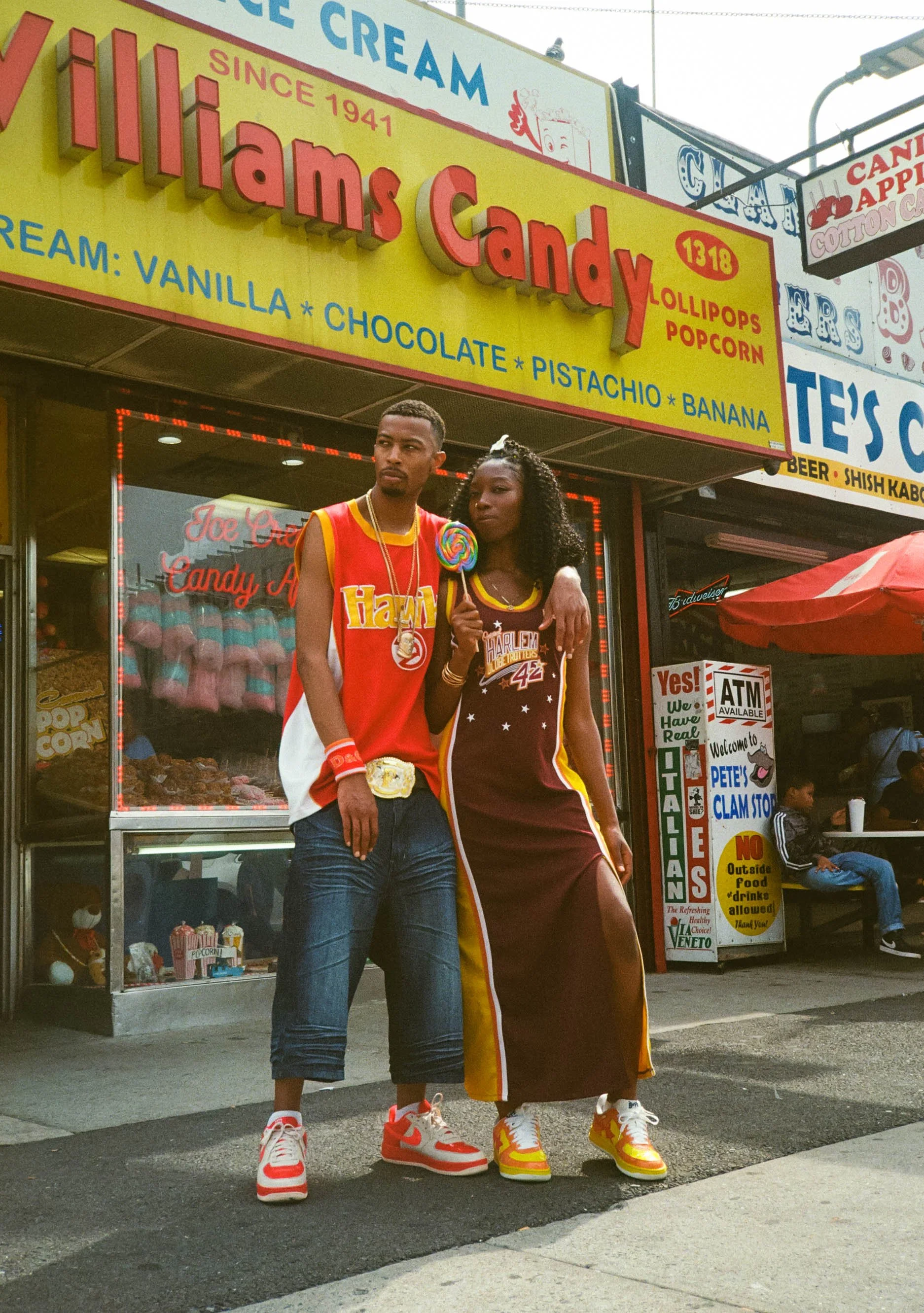 Two young adults standing in front of a candy shop labeled 'Williams Candy' with a yellow and red sign above. The male on the left is wearing a red basketball jersey, jeans, and sneakers, and has a championship belt around his waist. The female on th