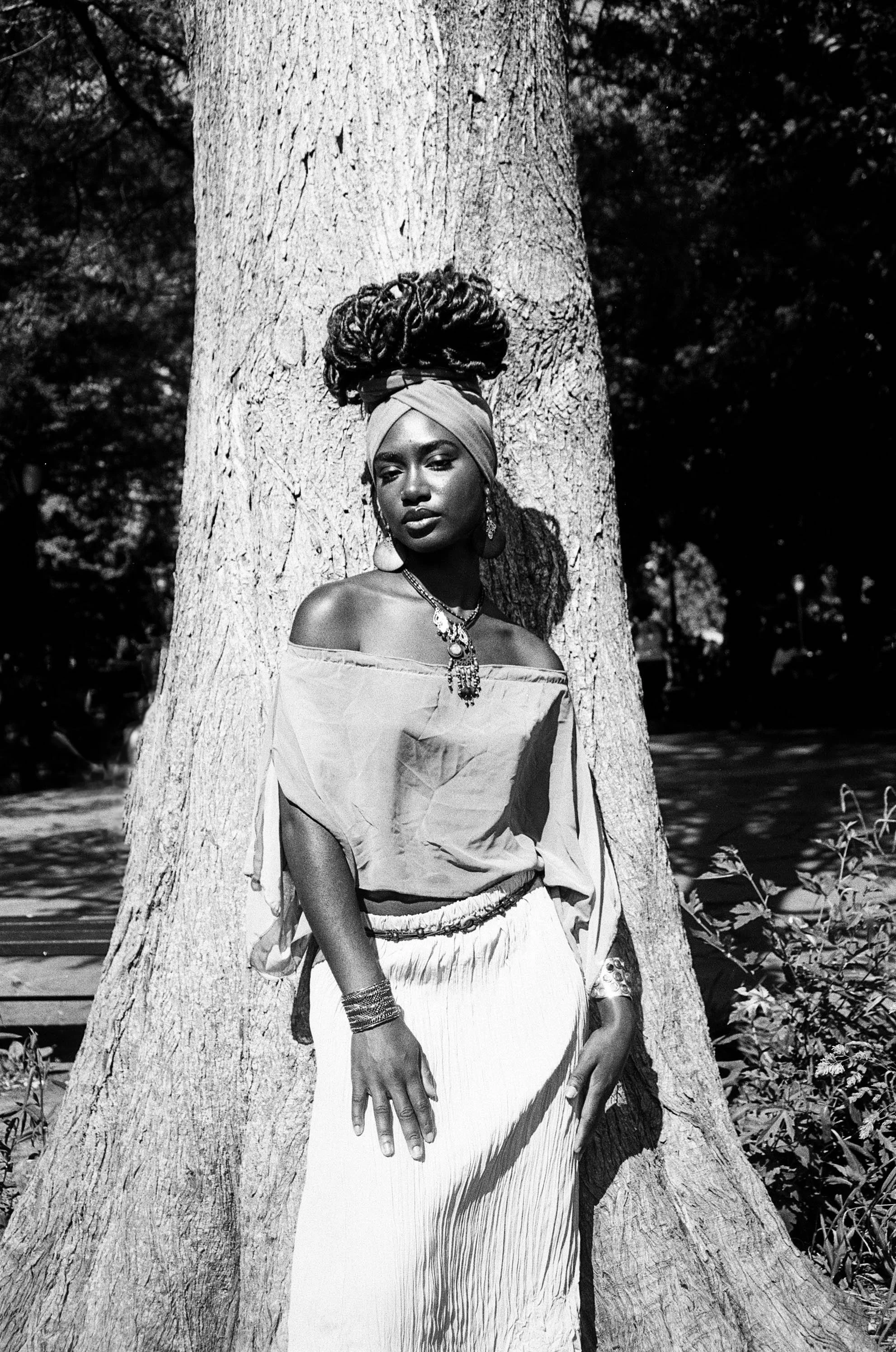 A Black woman stands outdoors behind a large tree, wearing a headwrap, earrings, necklaces, and a loose off-the-shoulder top and a long skirt, with a calm expression, in a natural setting with sunlight and shadows.