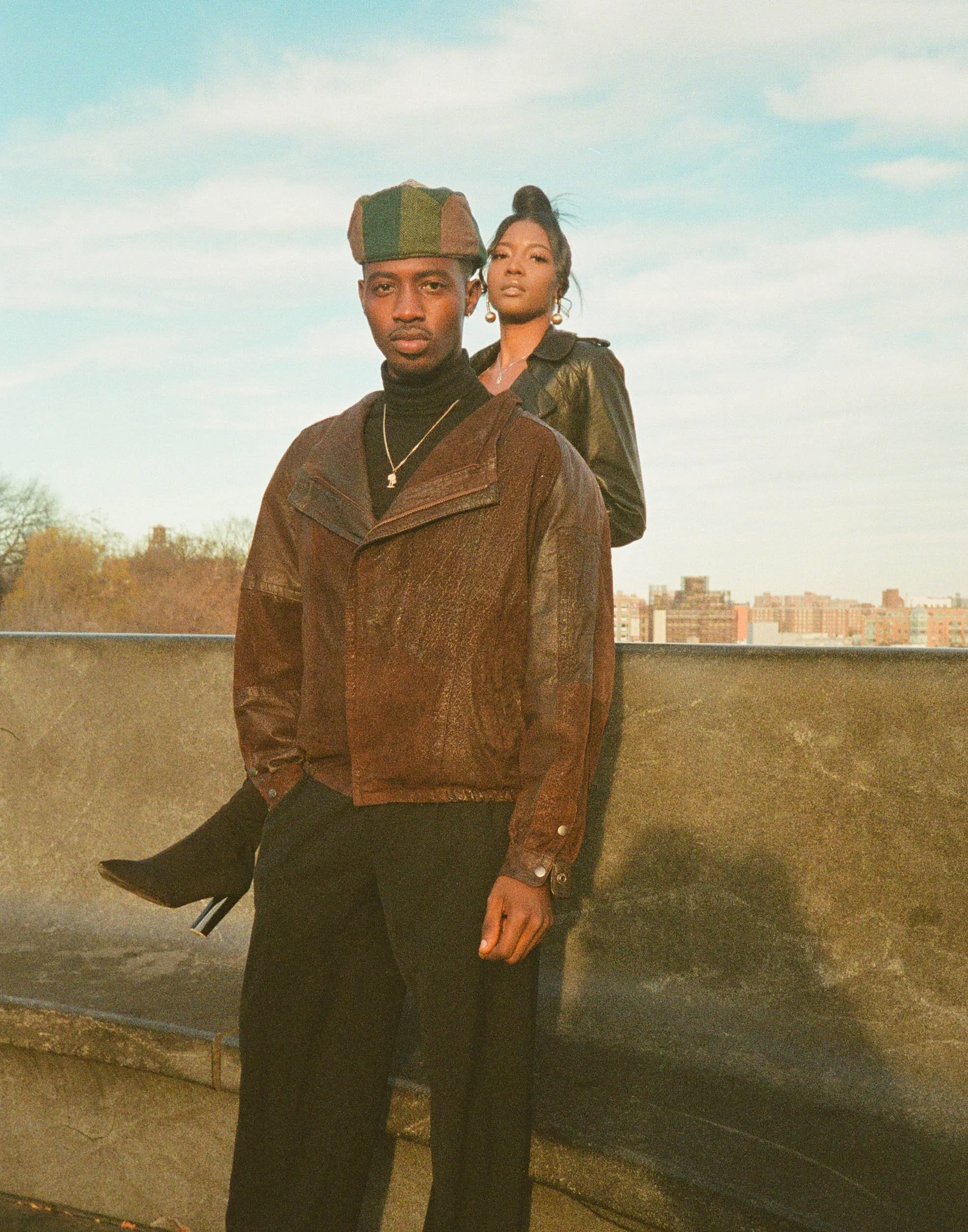 A man with dark skin wearing a multicolored beret, black turtleneck, and brown leather jacket standing in front of a cityscape during sunset, with a woman with dark skin and long hair styled in an updo standing behind him, both looking at the camera.