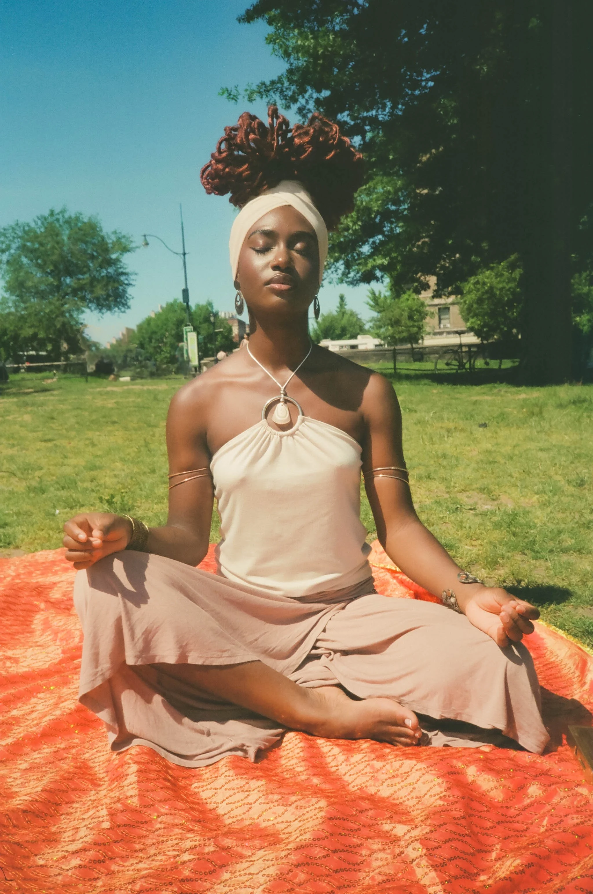 A woman meditating in a park on a bright, sunny day, sitting cross-legged with her eyes closed. She wears a beige headwrap, earrings, a necklace, and a flowing dress. She is seated on an orange patterned mat, surrounded by green trees and a clear blu