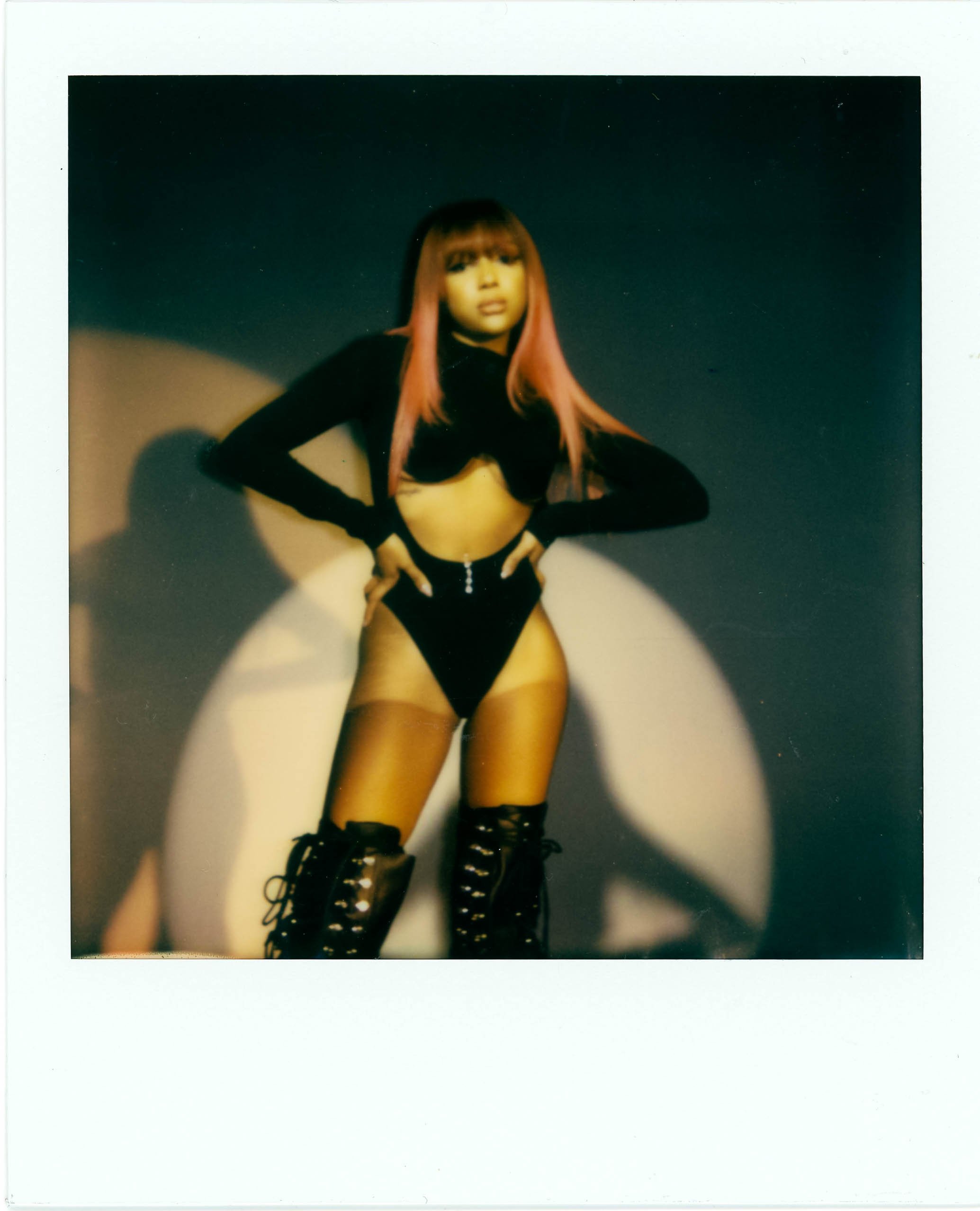A woman with pink hair wearing black lingerie and lace-up boots, standing with hands on hips in a spotlight against a dark background.