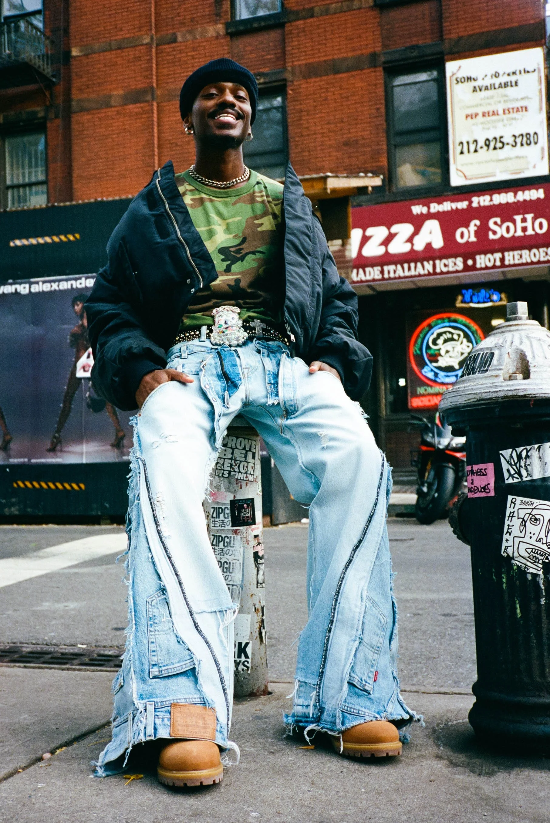 A young man wearing a black beanie, camouflage t-shirt, black jacket, and baggy ripped jeans standing on a city sidewalk near a fire hydrant and a pole covered in stickers. Behind him are brick buildings and signs for a pizza place and real estate.