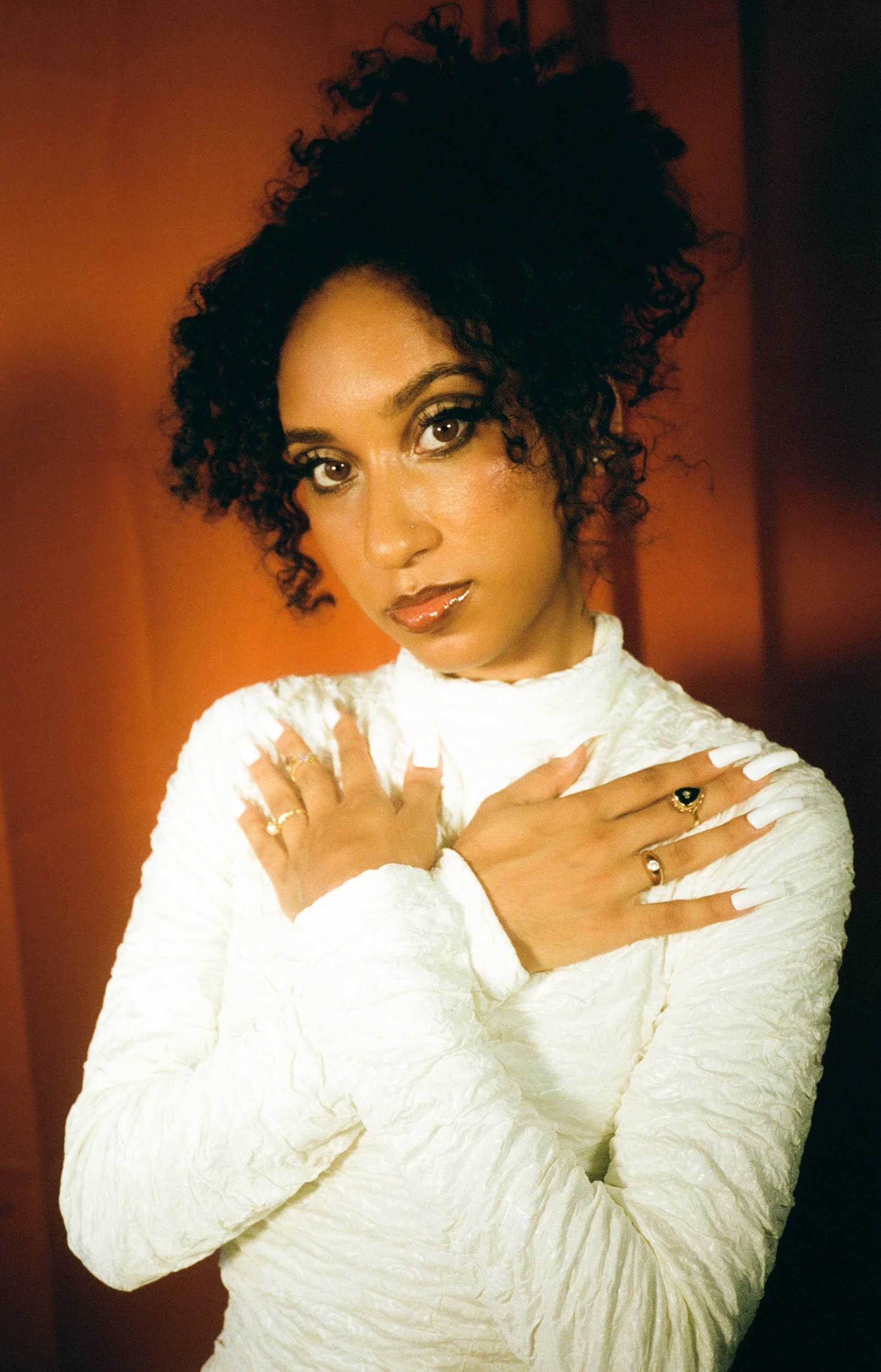 A woman with dark, curly hair and brown eyes, wearing a white textured long-sleeve top and multiple rings on her fingers, is standing with her arms crossed over her chest in front of an orange background.