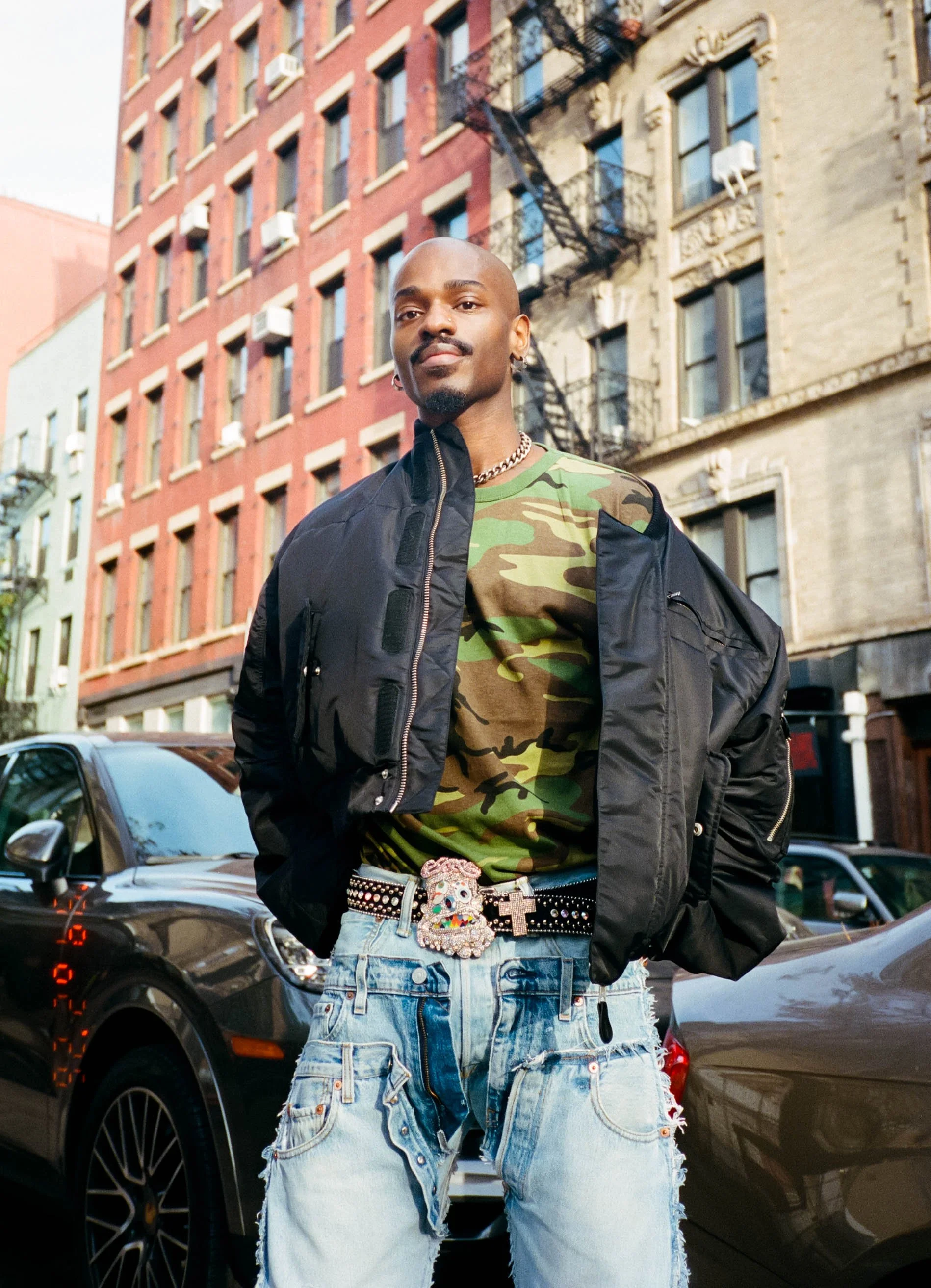 A man standing on a city street, wearing a black jacket over a green camouflage shirt, with distressed blue jeans, a colorful belt, and jewelry including a chain and earrings, with parked cars and classic brick buildings in the background.