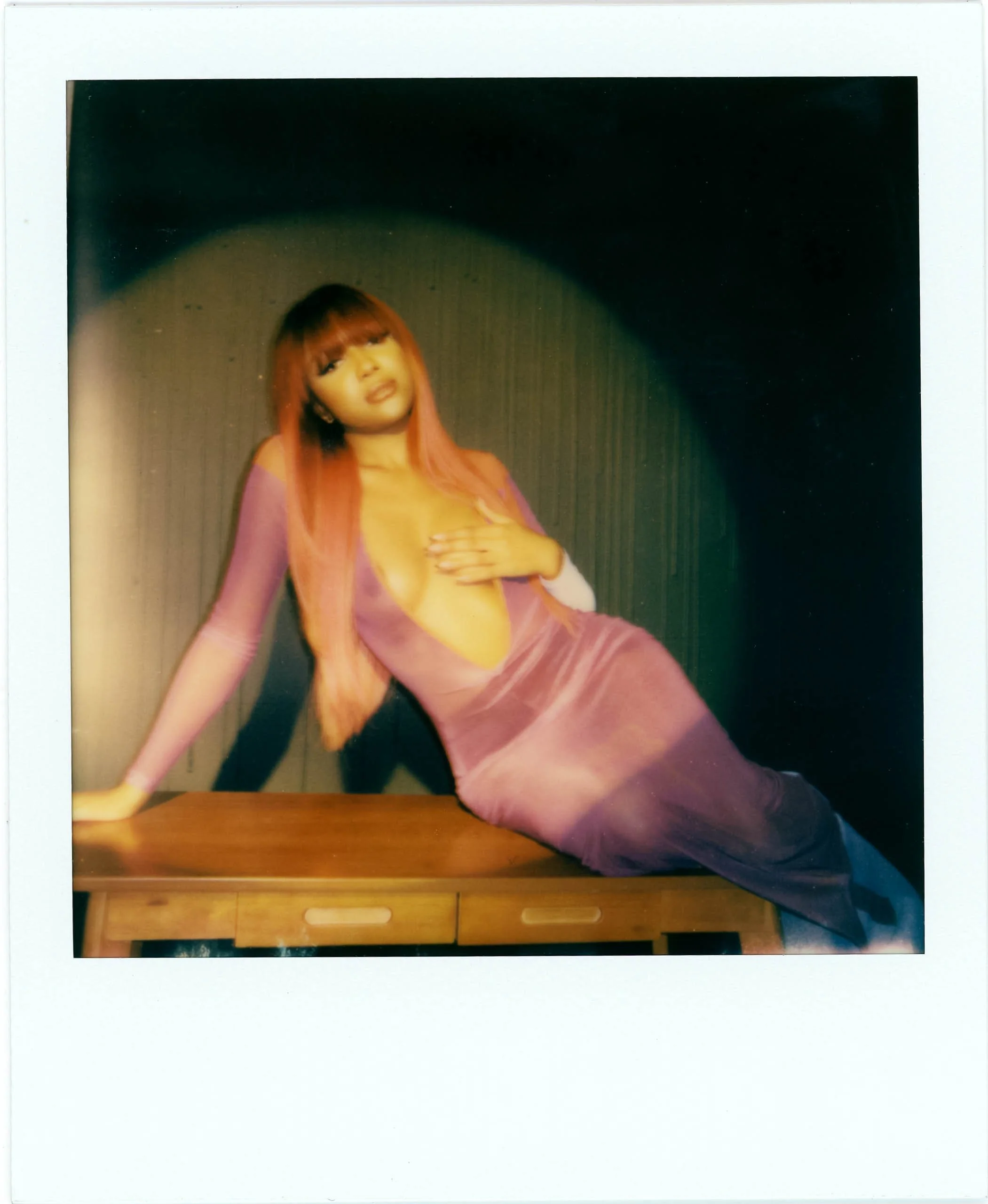 A woman with long pink hair wearing a pink and purple outfit, sitting on a wooden surface with a black background.