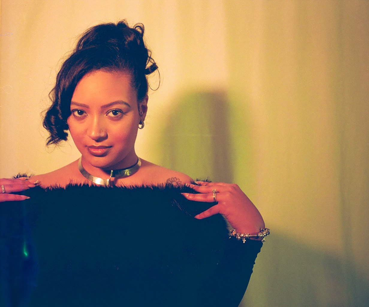 A woman with short, dark curly hair and light skin poses with her hands on her shoulders, looking directly at the camera with a slight smile, wearing a black fuzzy top and jewelry including a choker, rings, and bracelets, against a plain background.