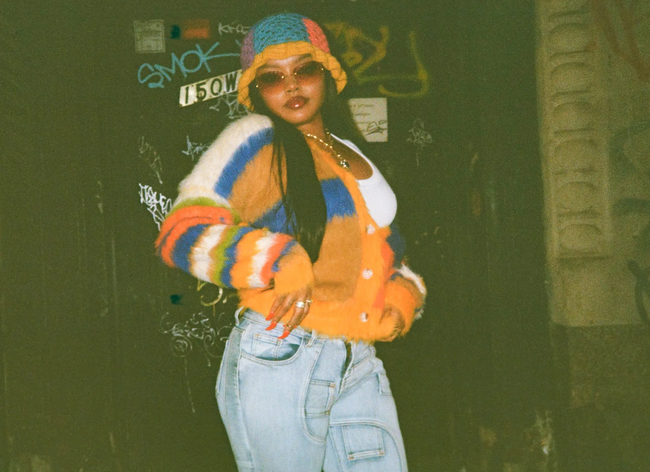 A woman wearing sunglasses, a colorful knitted hat, an orange and multicolored sweater, and light blue jeans, standing against a graffiti-covered wall.