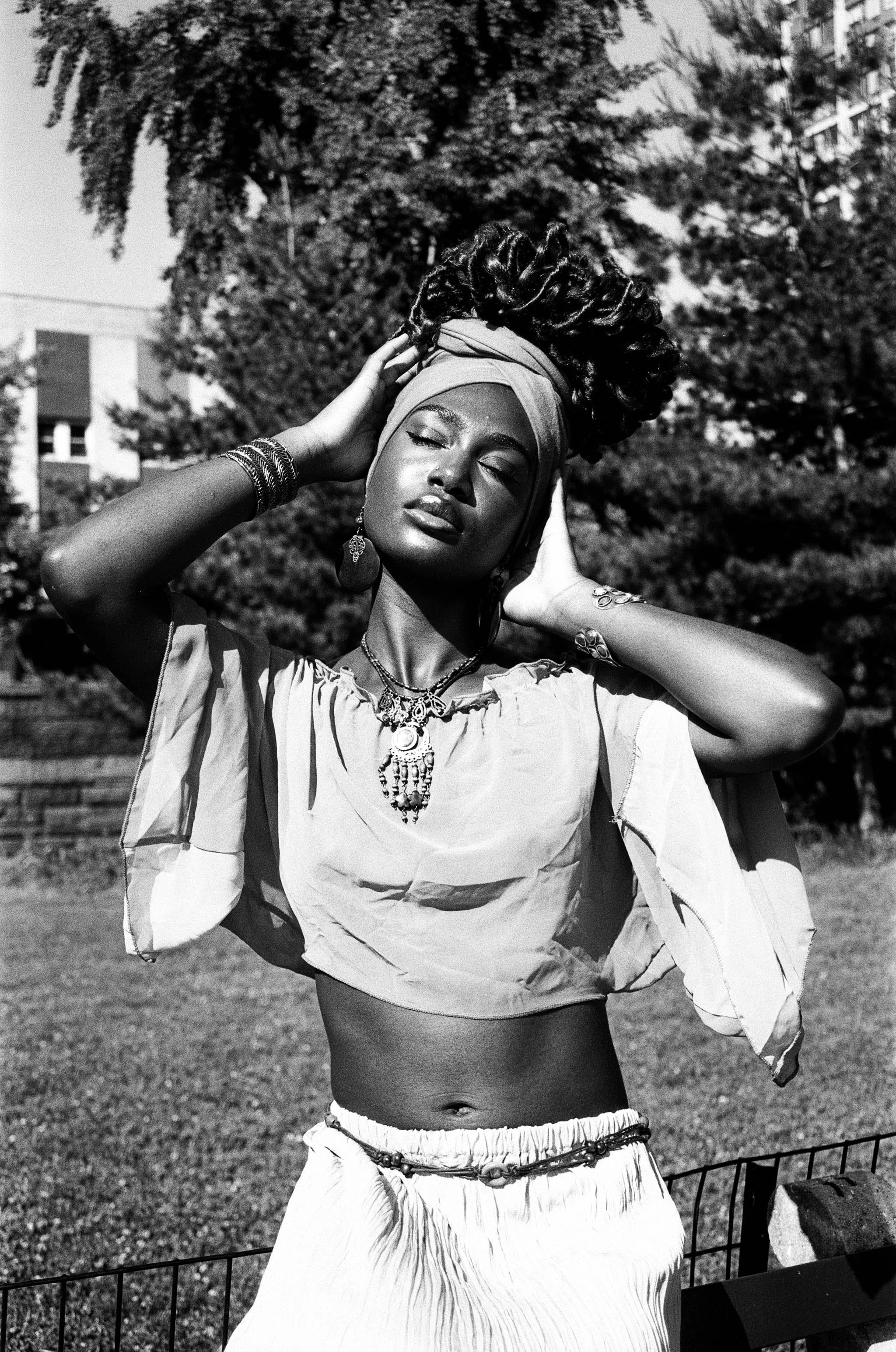 A woman with dark skin and closed eyes, dressed in a loose, light-colored top and skirt, wearing jewelry including earrings, necklaces, and bracelets, standing outdoors in front of trees and buildings, with her hands behind her head and her hair styl