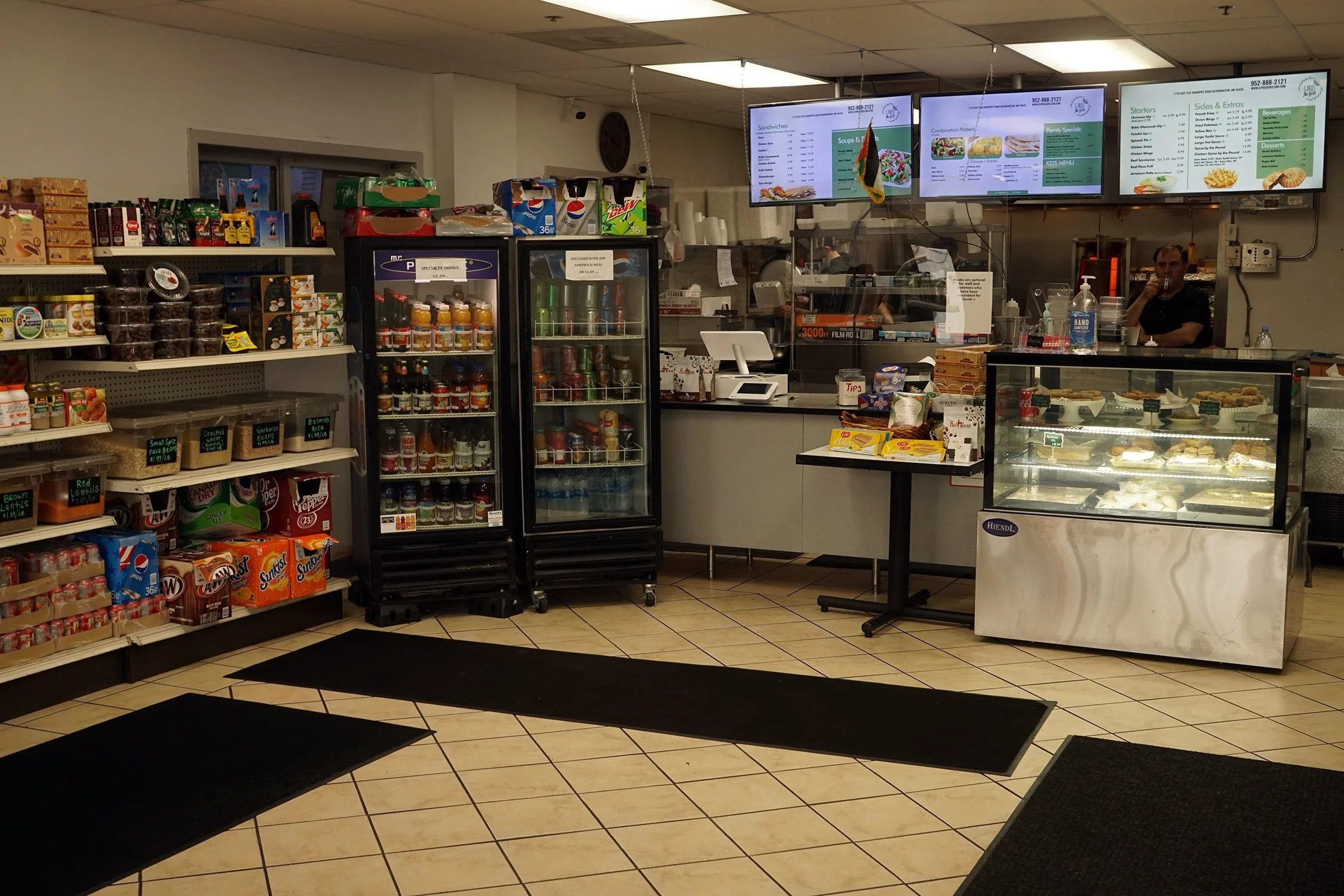 Inside a small cafe or deli with a counter, digital menu screens above, a display case of desserts, and retail shelves with snacks and drinks.
